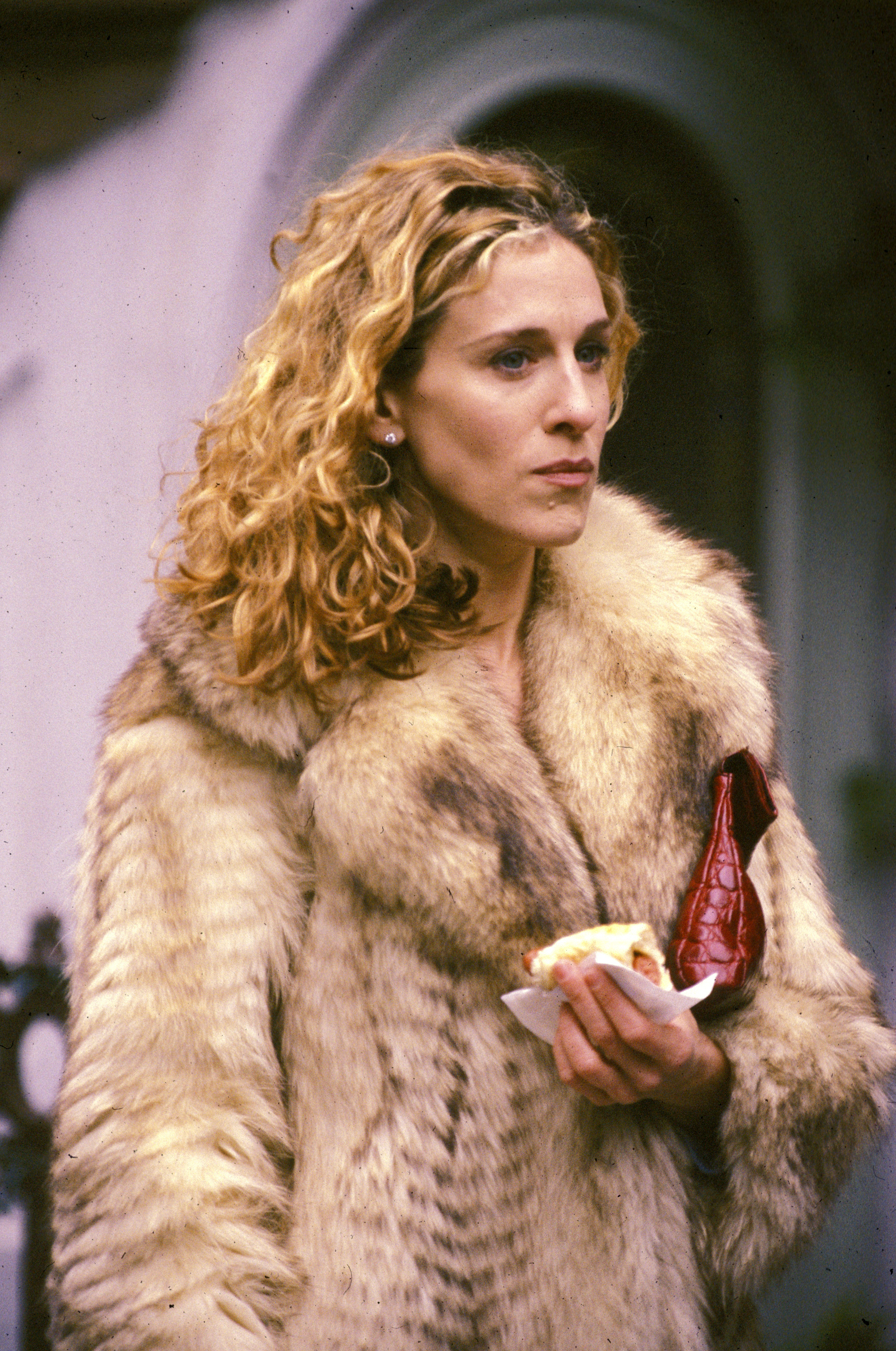 Sarah Jessica Parker, wearing a fur coat, holds a snack in one hand and appears to be standing outdoors