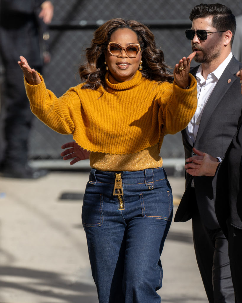 Oprah Winfrey, wearing a stylish turtleneck sweater and high-waisted jeans, gesturing with open arms while walking with a man in a suit and sunglasses