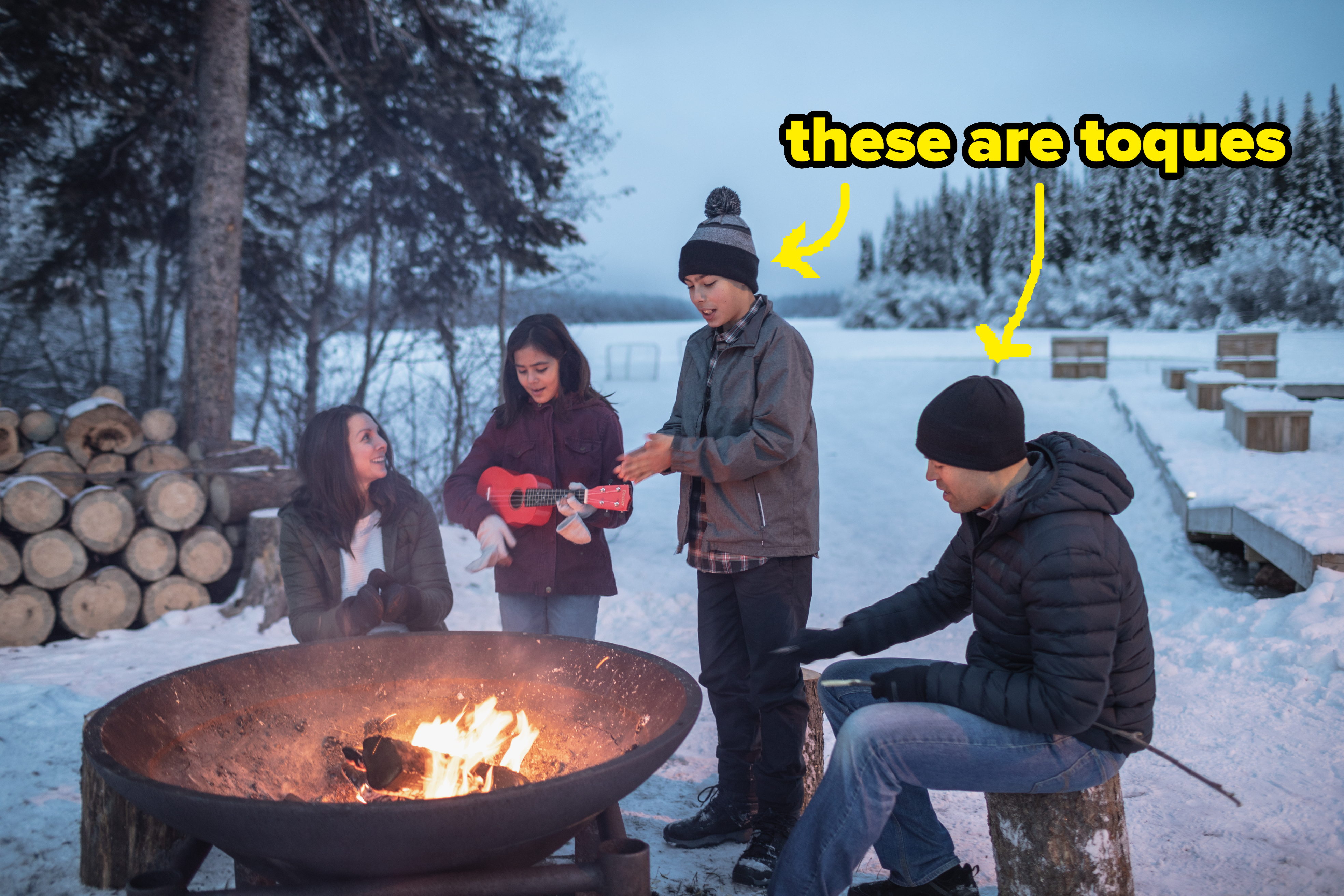 A group of four friends gather around a large outdoor fire pit in the snow, with one person playing a ukulele
