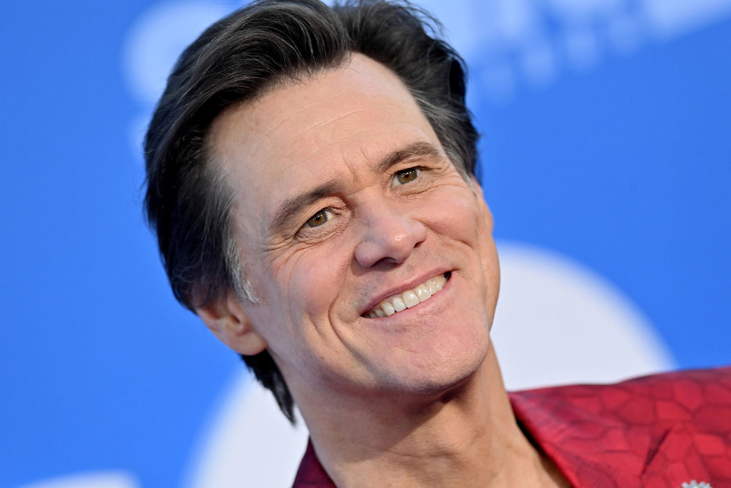Jim Carrey smiles, wearing a red, patterned jacket with a blurred blue background