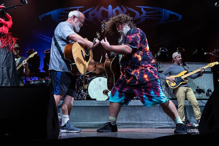 Jack Black and Kyle Gass of Tenacious D perform energetically on stage, both playing guitars. A bassist and drummer are also visible in the background
