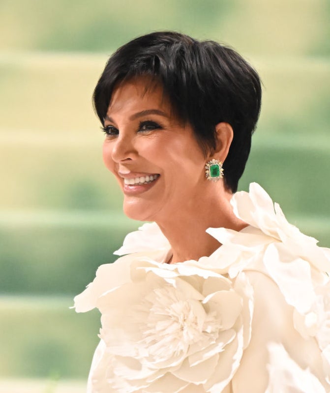 Kris Jenner smiles while wearing a floral-themed top with large, ruffled flower details on the shoulders and sleeves. She has square earrings