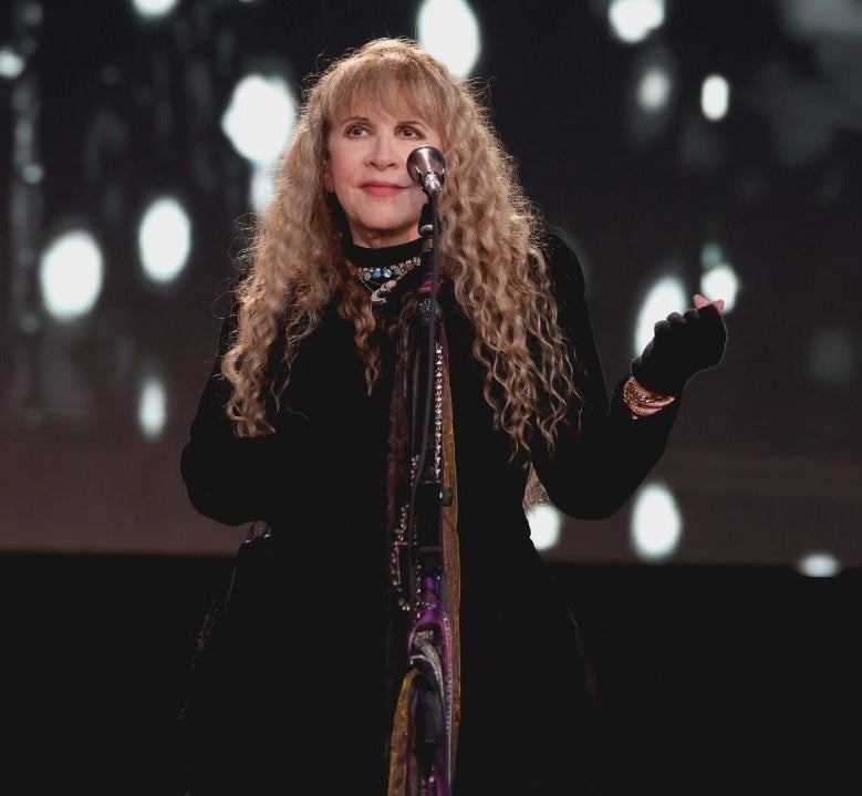 Stevie Nicks performing on stage in a dark outfit and gloves, with a microphone in front of her