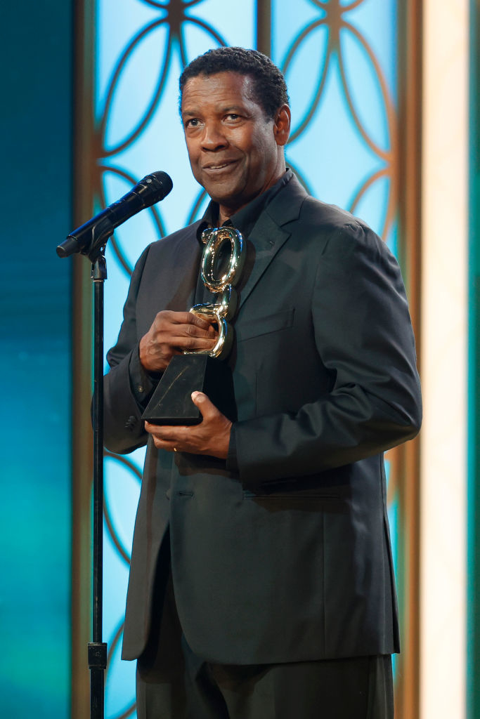 Denzel Washington stands at a microphone holding an award statue, dressed in a formal dark suit, with a backdrop featuring geometric patterns