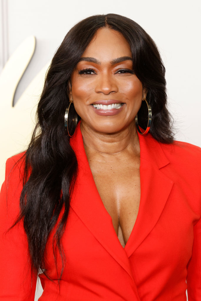 Angela Bassett smiling, wearing a stylish, deep V-neck red blazer with large hoop earrings