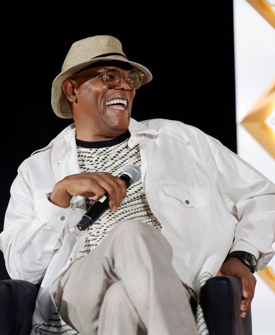 Samuel L. Jackson smiles and holds a microphone while seated onstage, wearing a hat, glasses, and casual clothing
