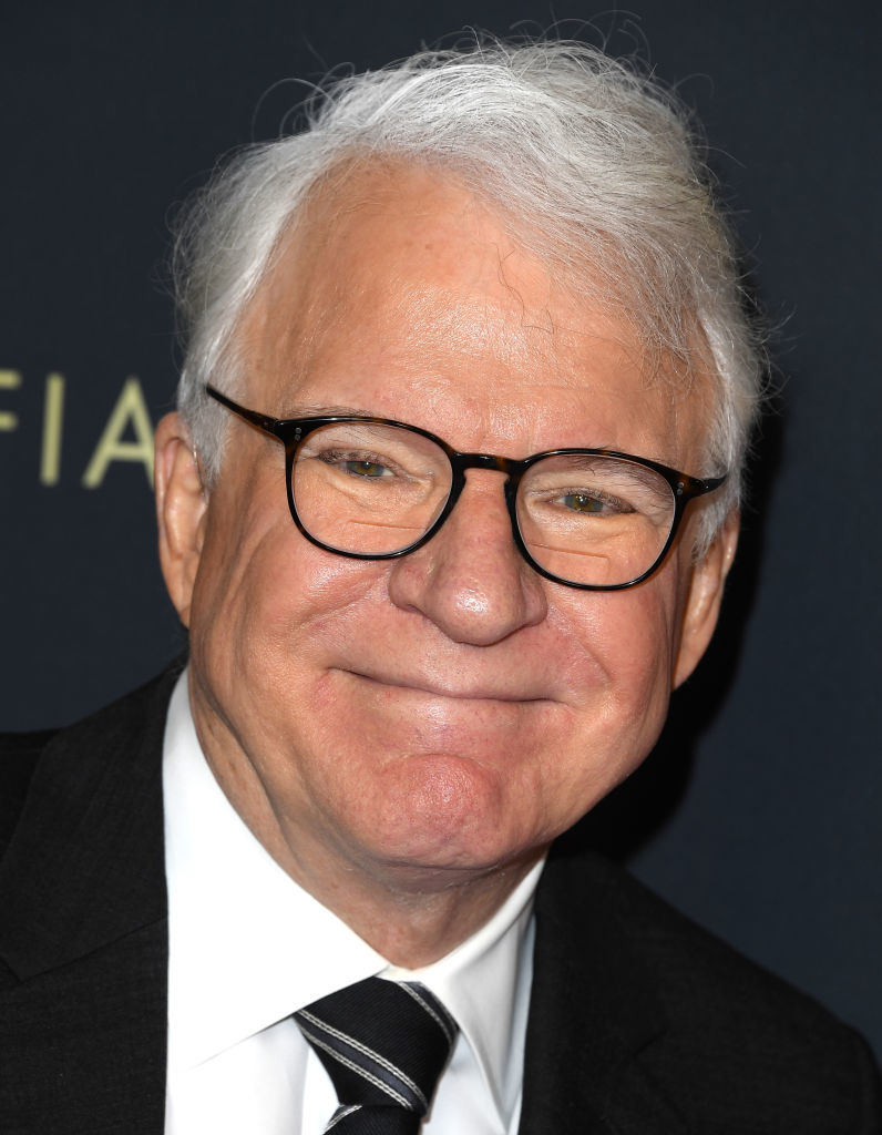 Steve Martin smiling, wearing a dark suit, white shirt, and striped tie