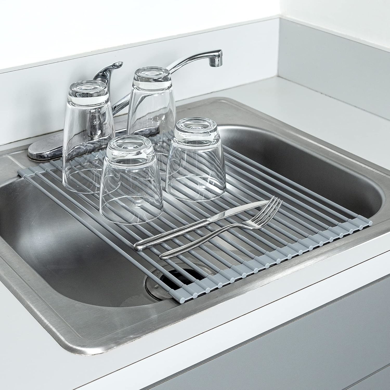 Clean dishes and cutlery dry on an over-the-sink dish rack, including four glasses, a fork, and a knife