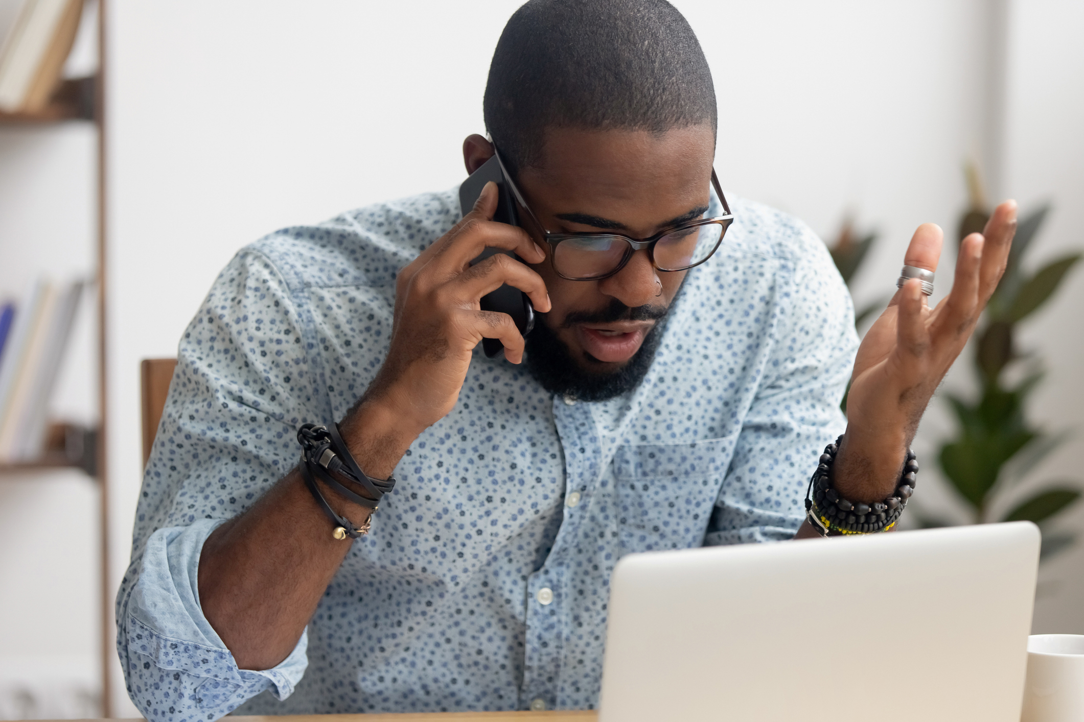 A man wearing a patterned shirt and glasses is on a phone call, gesturing with one hand while looking at a laptop screen