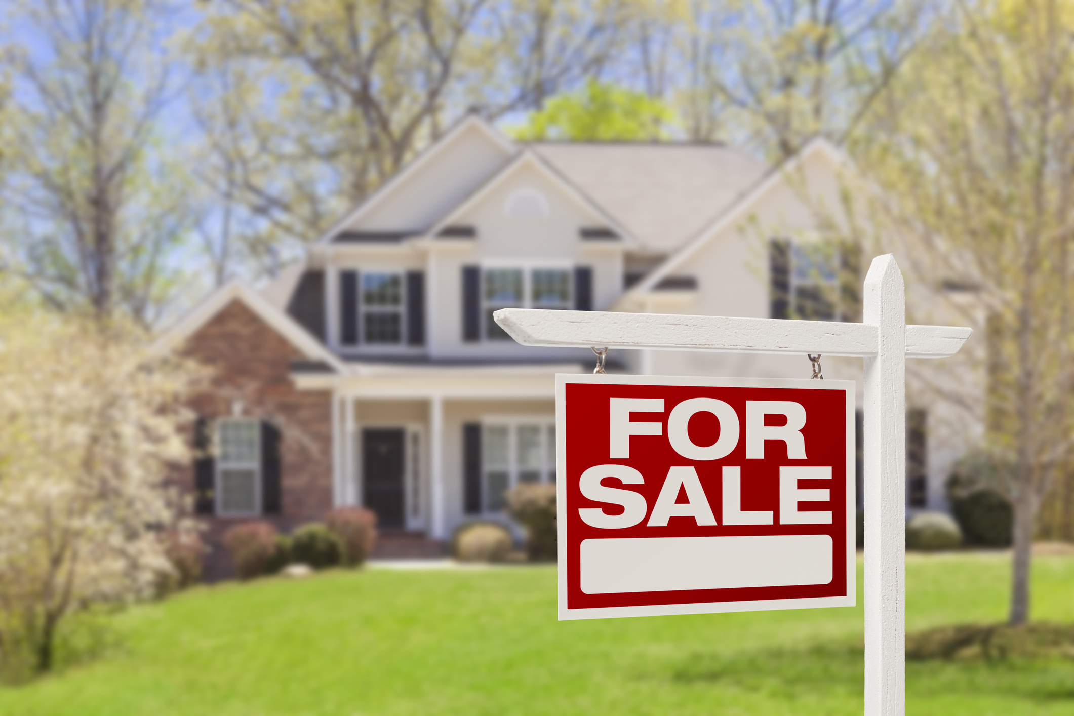 A large house with a "For Sale" sign in the front yard. Trees and a well-maintained lawn surround the house