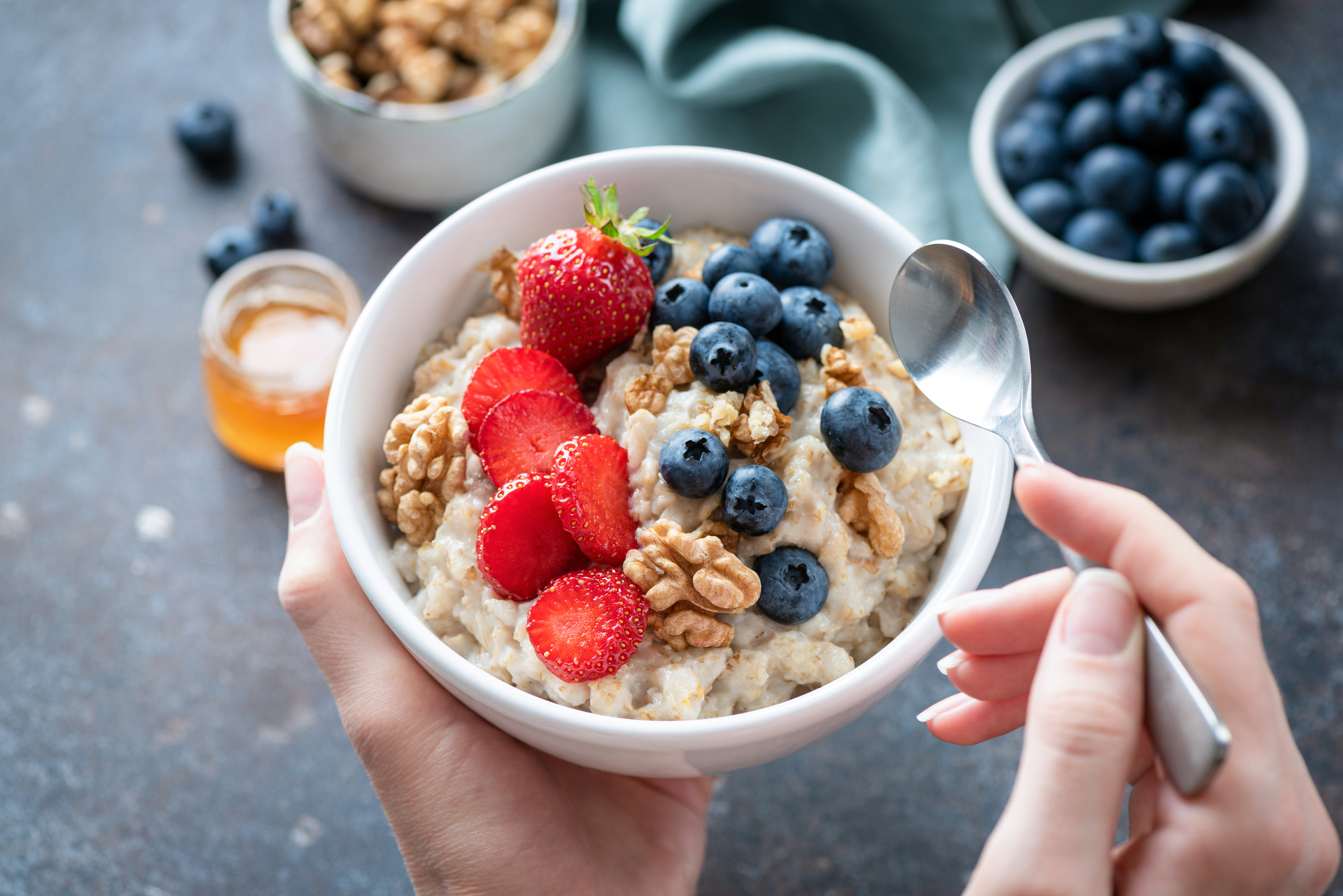 A bowl of oatmeal topped with strawberries, blueberries, and walnuts being held with a spoon ready to eat. Honey and additional blueberries are in small bowls nearby