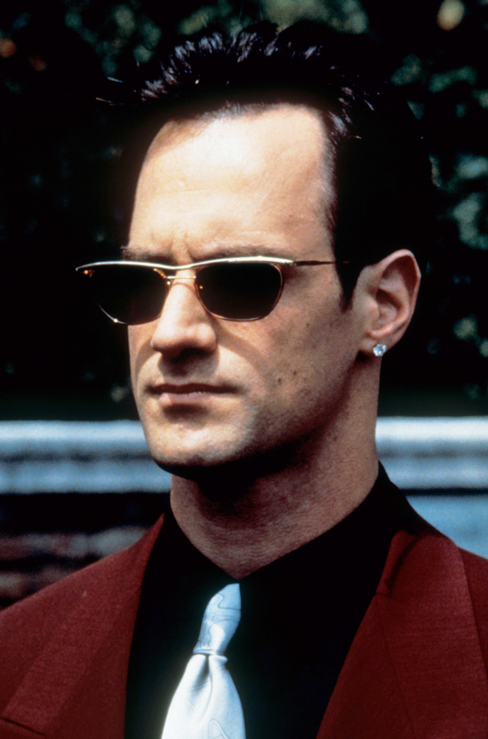Christopher Meloni wearing sunglasses, a dark red blazer, and a light-colored tie, looking straight ahead with a serious expression