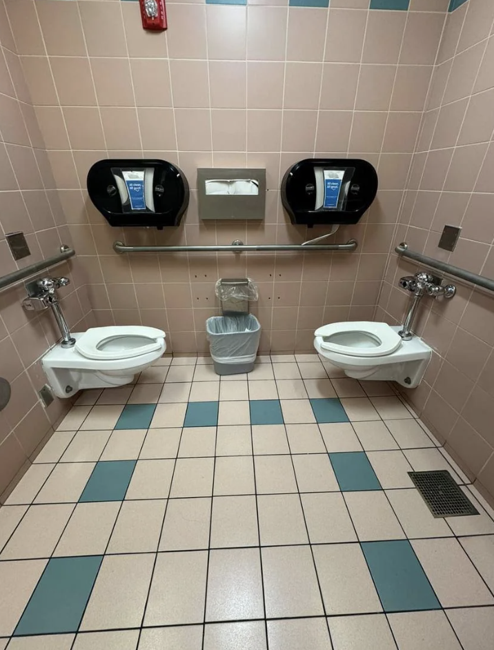 Two side-by-side toilet seats in a public restroom, each with a toilet paper dispenser above. A waste bin is placed between the toilets