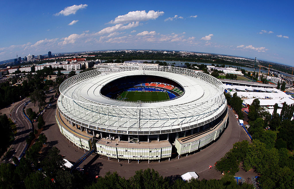 Aerial view of a large, circular stadium with a field and seating inside, surrounded by trees and city buildings in the distance