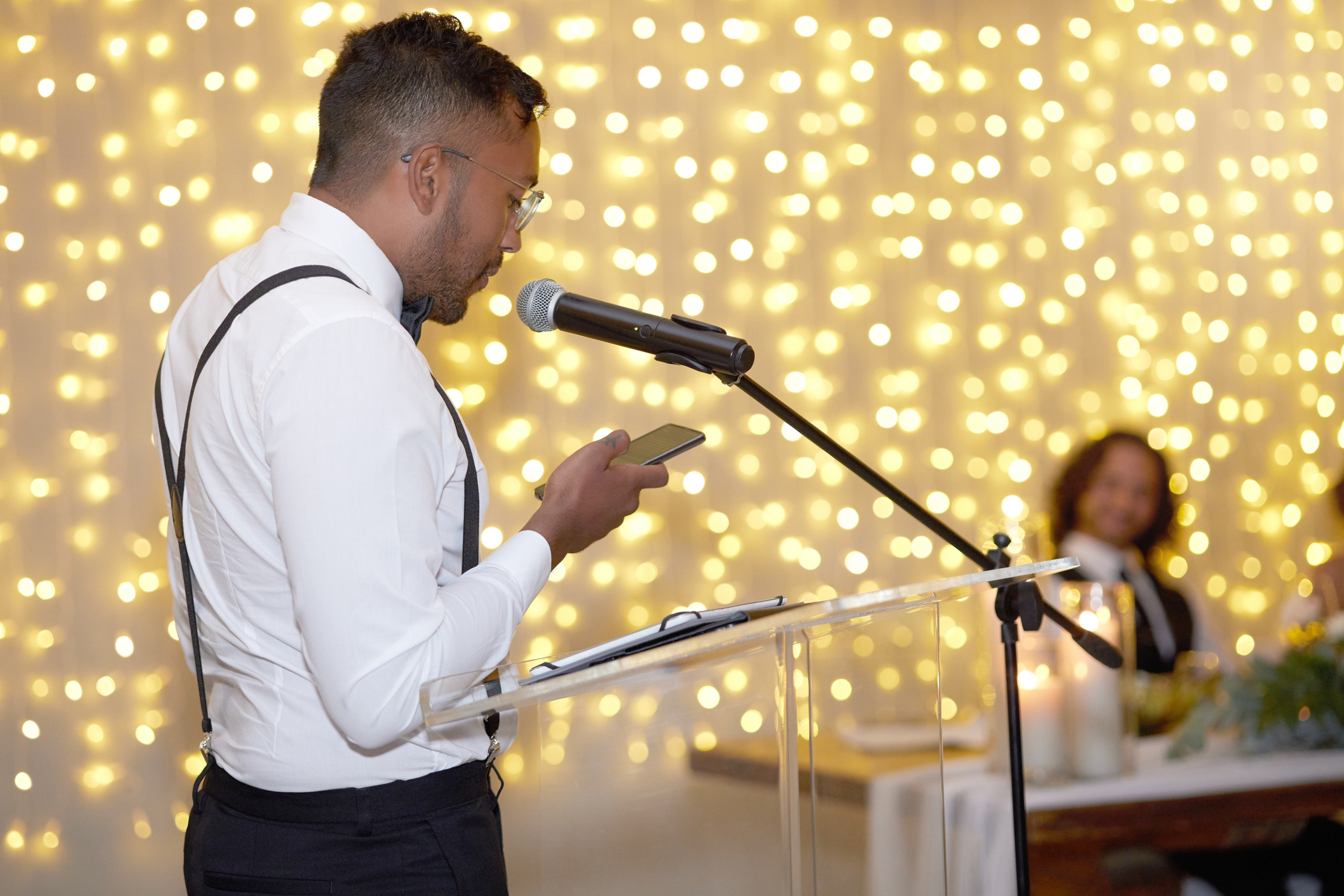 A man wearing a white shirt and black suspenders speaks into a microphone at a podium; a person in the background listens smiling