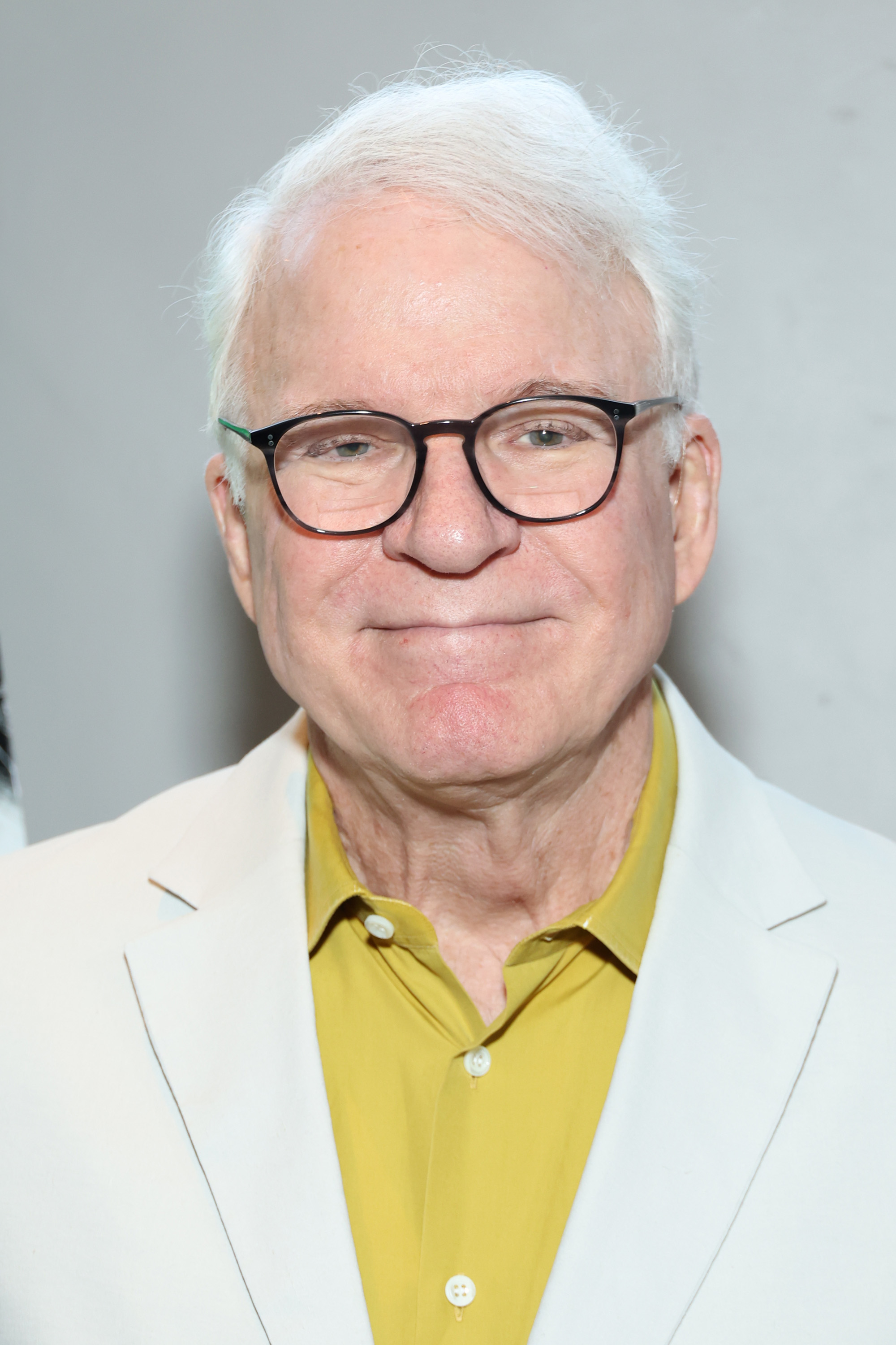 Steve Martin smiling, wearing glasses, a light blazer, and a yellow shirt