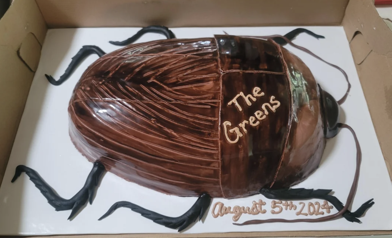 Cockroach-shaped cake with icing text reading "The Greens" and "August 5th, 2024" on a white cake board