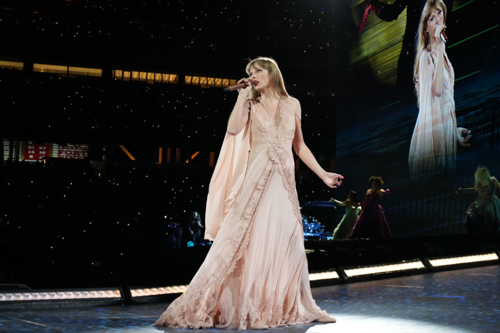 Taylor Swift performs on stage in an elegant, flowing dress during her concert, with a large screen displaying her image in the background