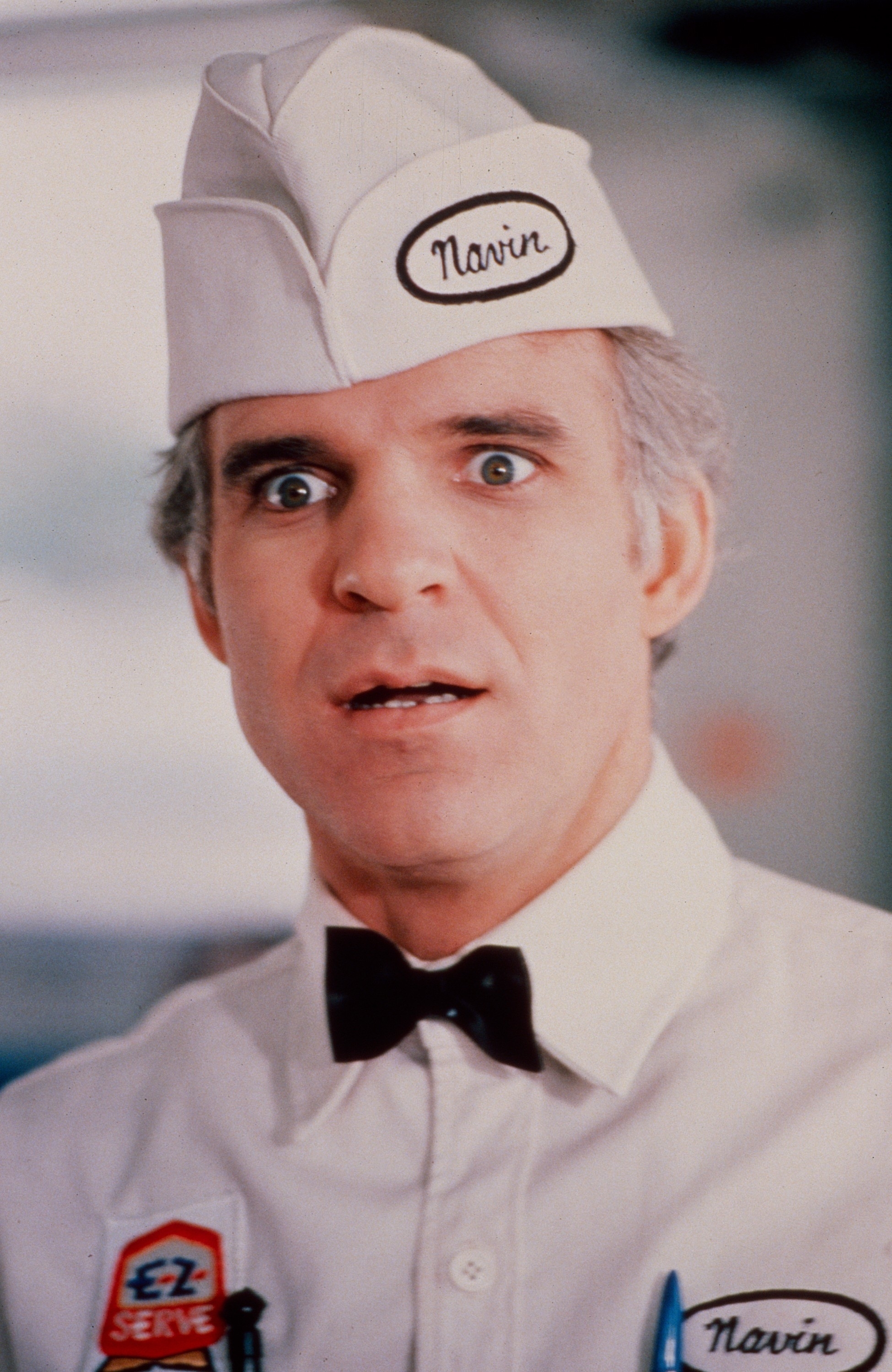 Steve Martin dressed as a soda jerk, wearing a white uniform with a bow tie and cap labeled "Navin", looking surprised