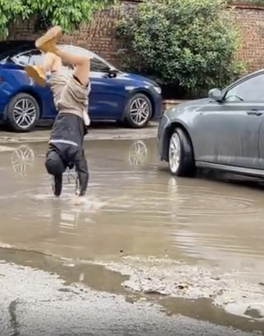 A person is doing a handstand in a large puddle on a street lined with parked cars, splashing water around