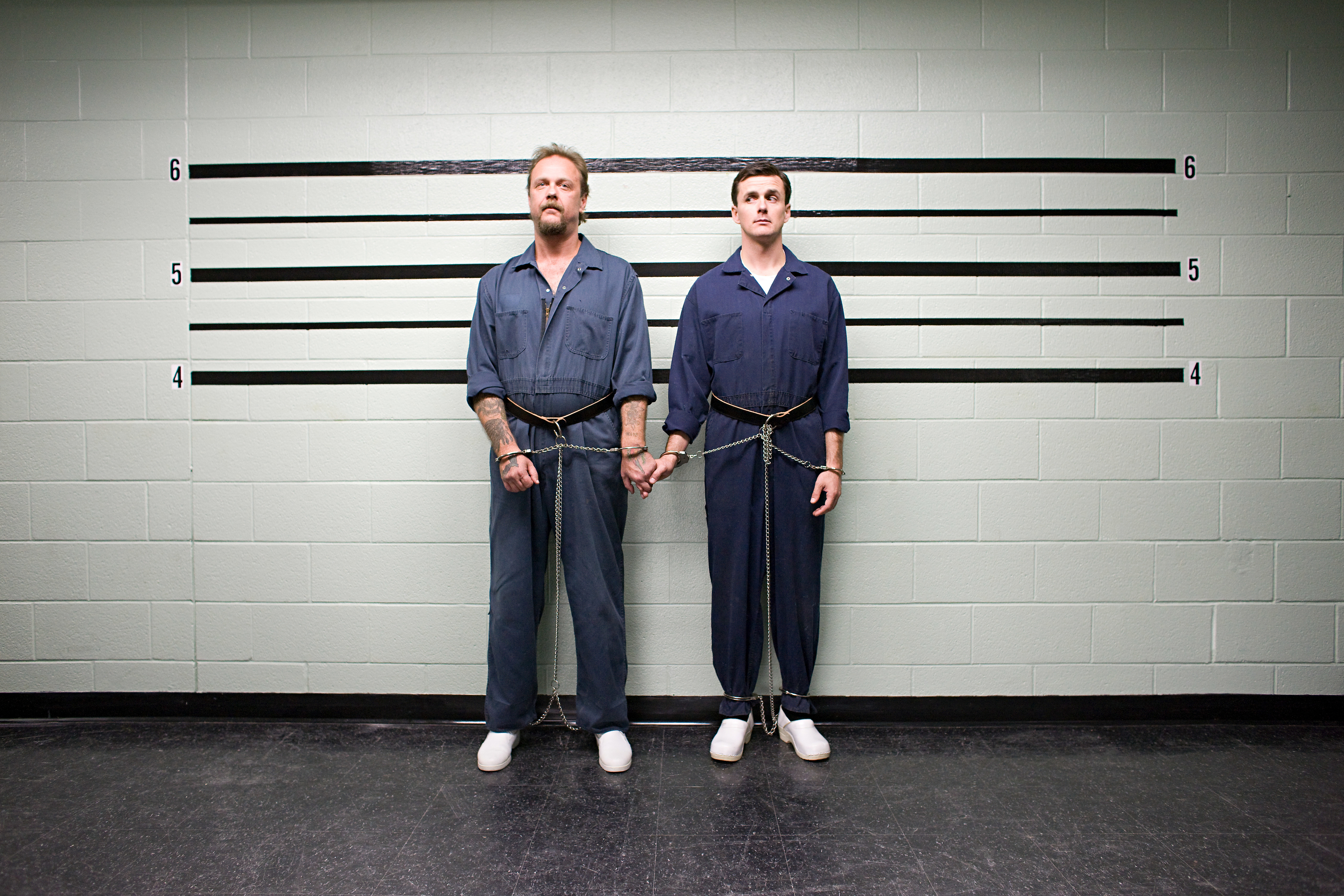 Two individuals in prison uniforms stand handcuffed and shackled side by side in front of a height chart. No names provided