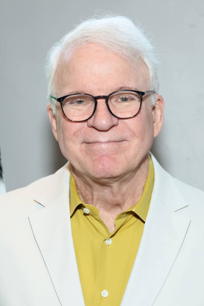 Steve Martin smiles, wearing glasses, a yellow shirt, and a light-colored jacket