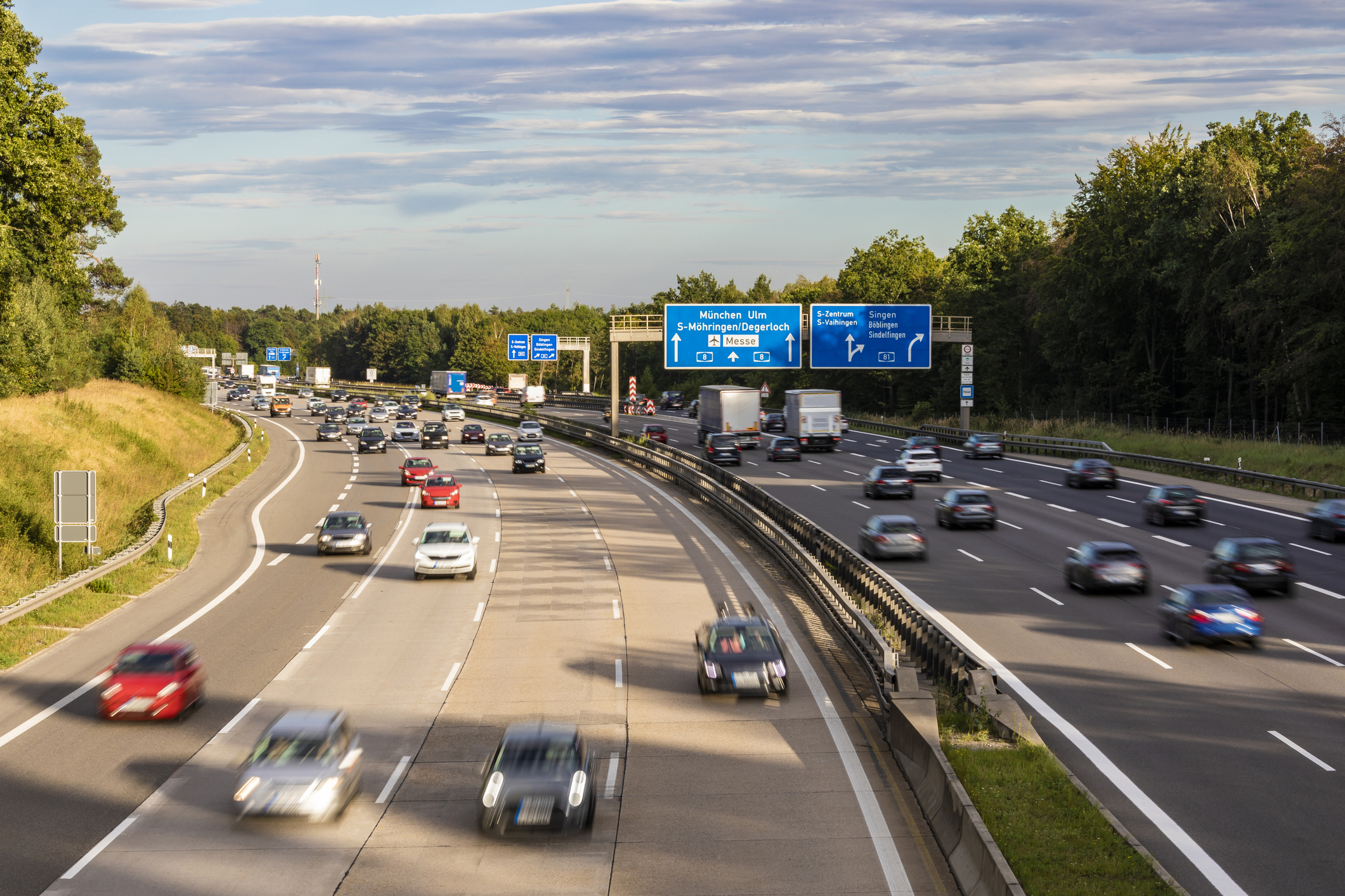 A busy multi-lane highway with various cars in motion. Road signs indicate directions to Munich, Ulm, and other locations. Trees line both sides of the highway