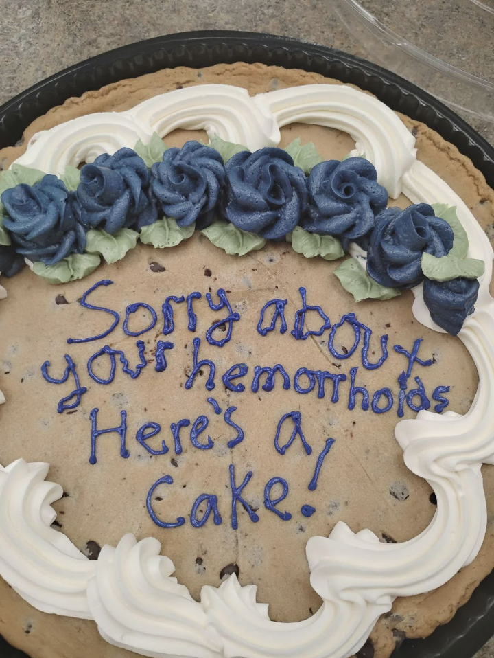 A large cookie cake with blue and white icing reads, "Sorry about your hemorrhoids Here's a cake!"