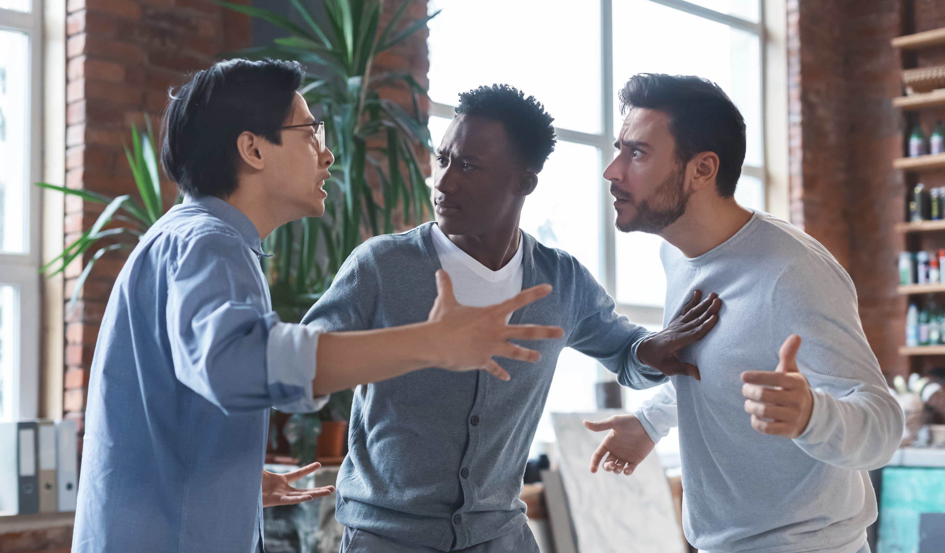 Three men are engaged in an animated discussion inside a modern office with exposed brick walls and large windows. No names are provided