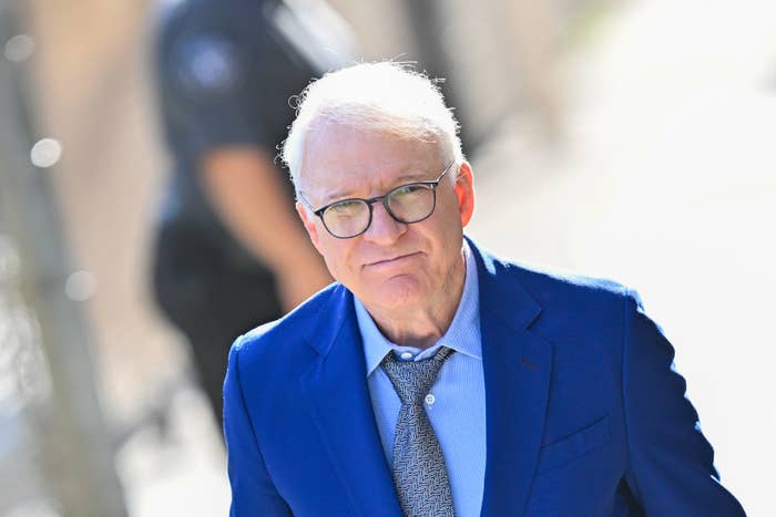 Steve Martin, in a suit with a tie, walks outside during daytime with neutral expression. Background shows a blurred person and a fence