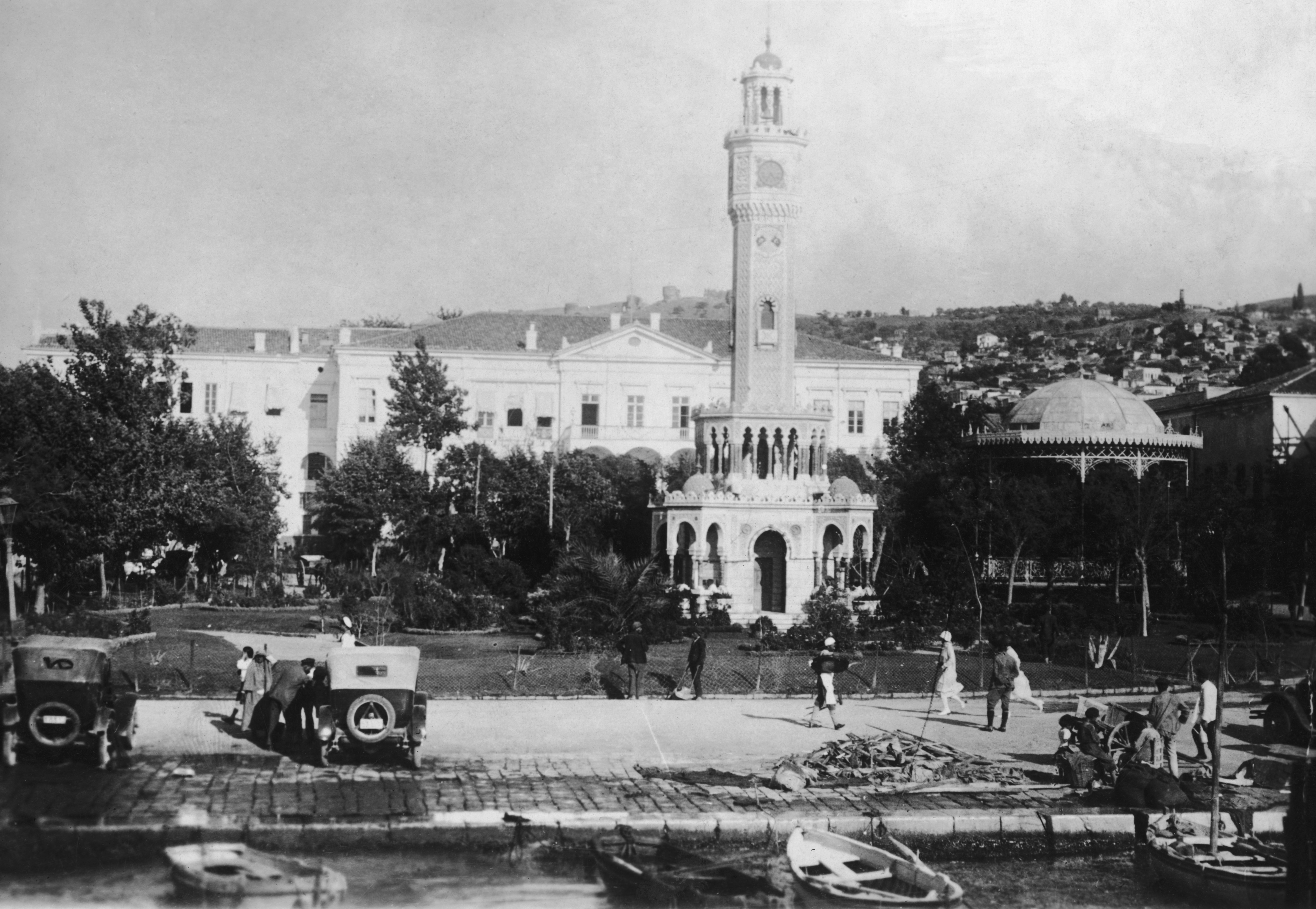 Historic photo of Izmir's Konak Square with the Clock Tower, people walking, and old vehicles parked. The background features the government building and trees in eastern Turkey, circa 1920.