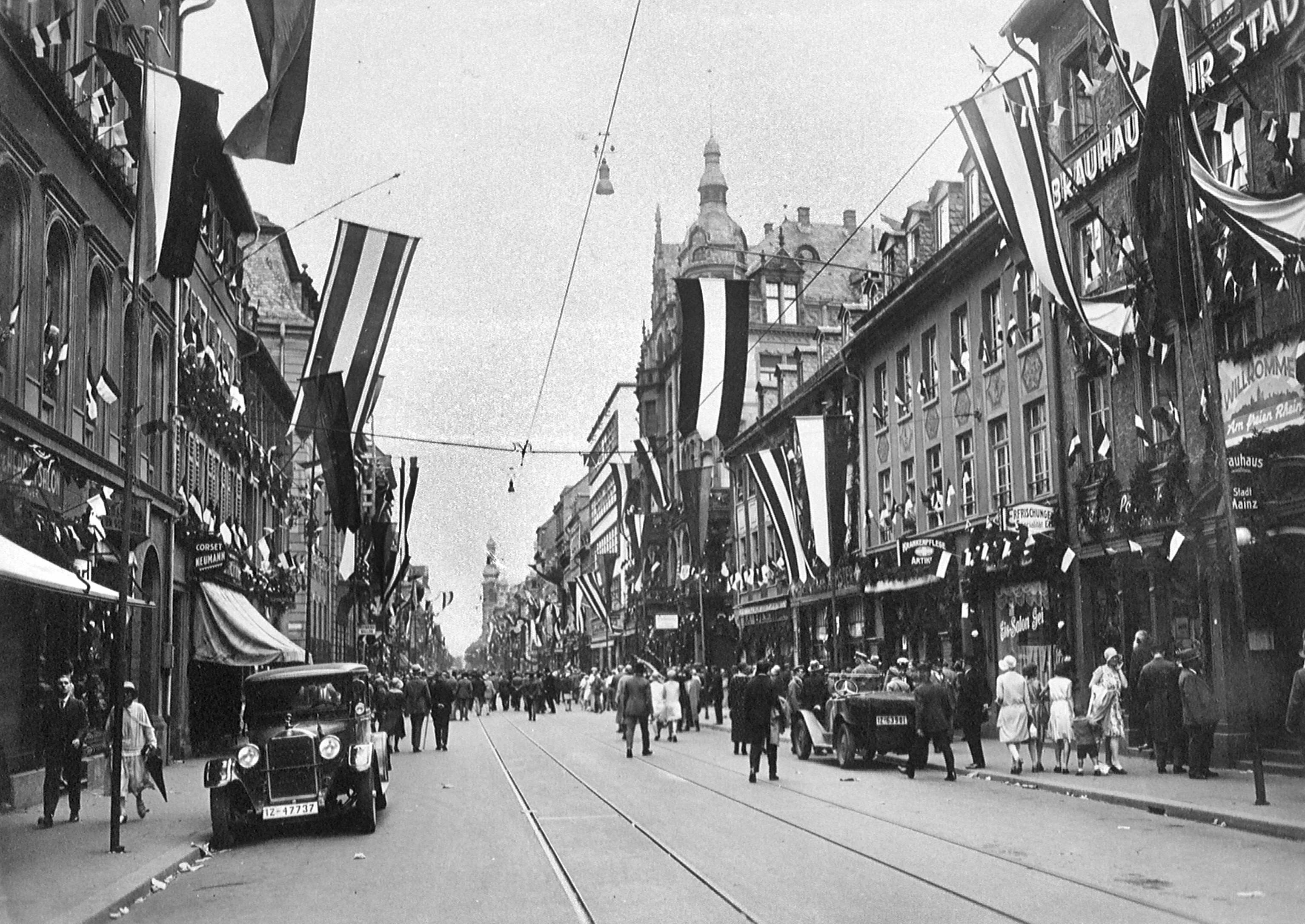 Historic main street scene with people walking, flags hanging from buildings, and period cars from Mainz Germany, circa 1920