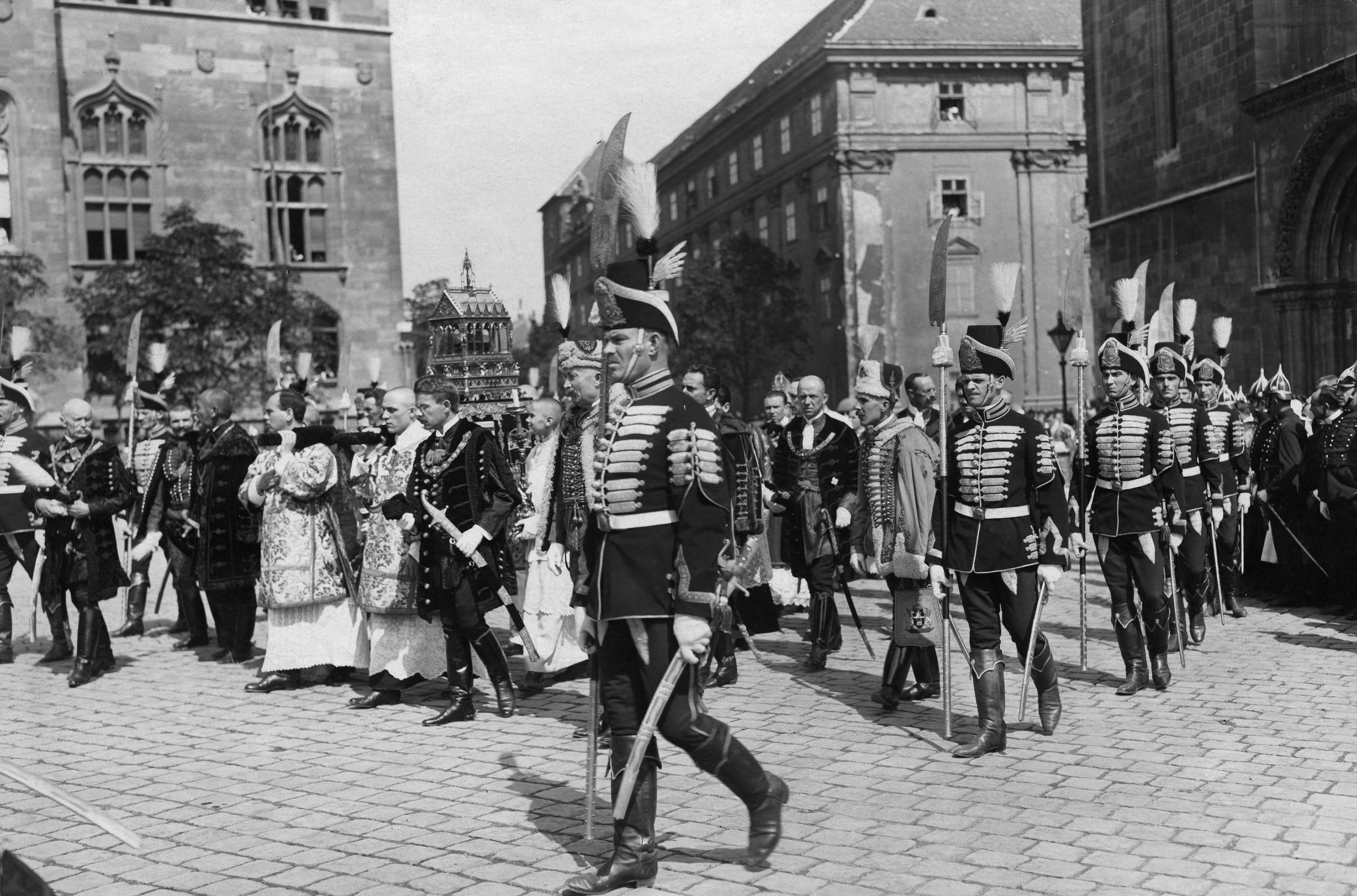 A historical procession featuring soldiers in ceremonial uniforms with swords, clergymen in ornate robes, and several onlookers, all gathered in a paved courtyard at St. Stefan's Day in Budapest, Hungary