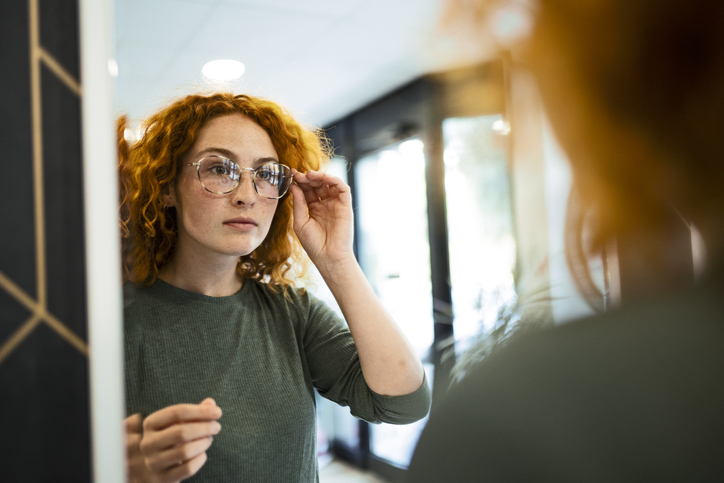A person with curly hair adjusts their glasses while looking into a mirror in a well-lit room
