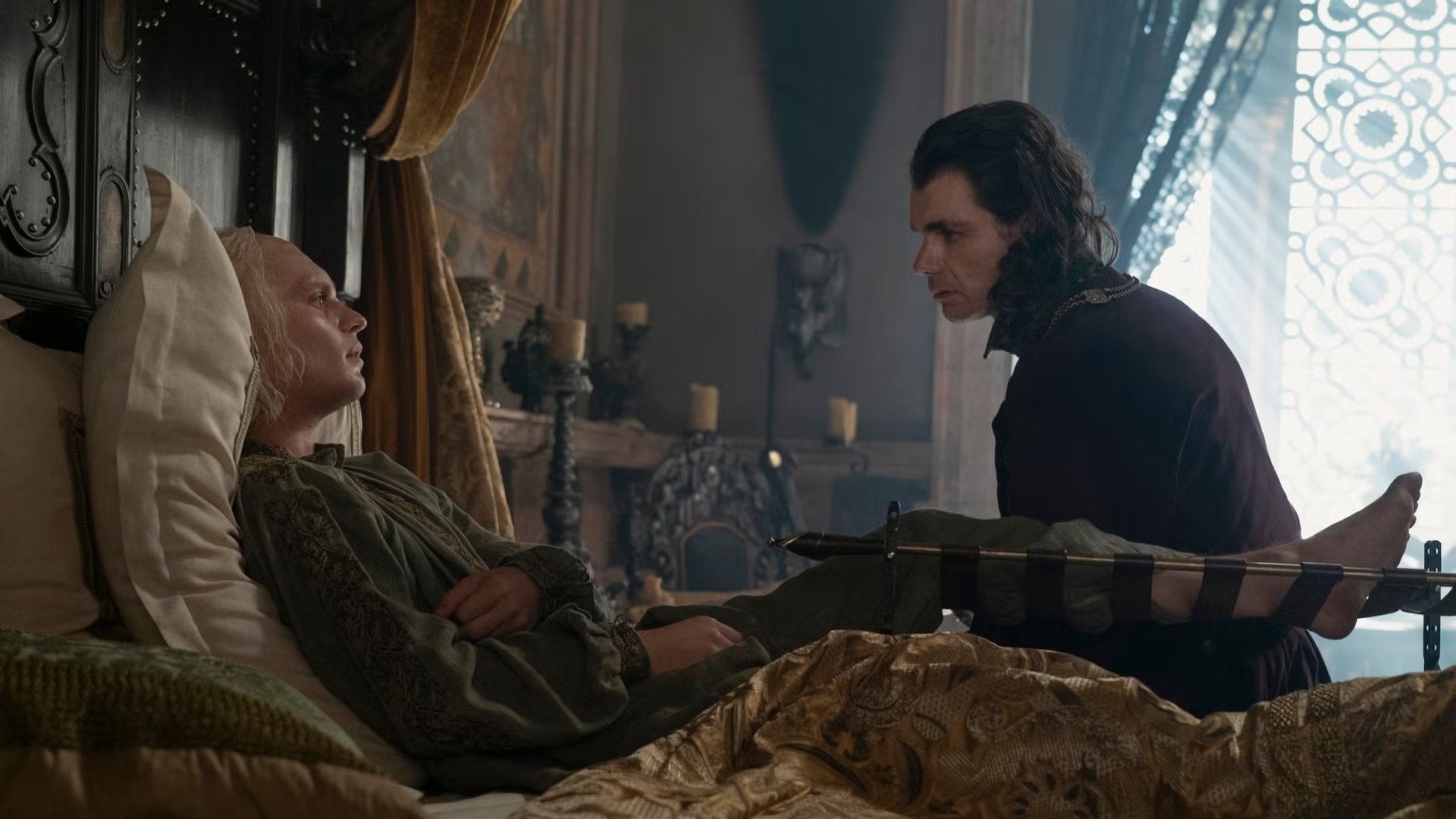 Viserys Targaryen yace en la cama, Rhaenyra Targaryen lo mira con preocupación. Escena interior ambientada en la serie basada en "Juego de Tronos"