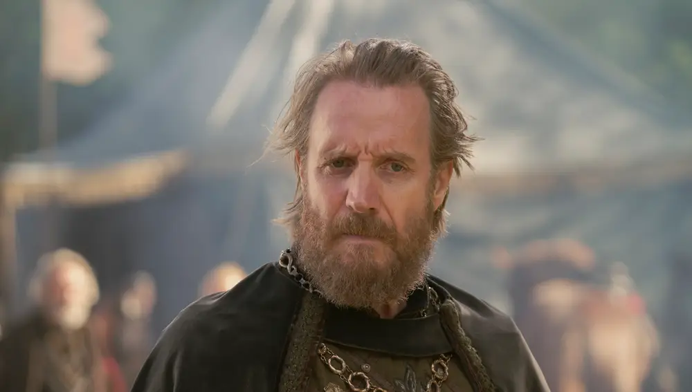 No sé quién es esta persona. Hombre con barba y cabello canoso se encuentra en lo que parece ser un ambiente medieval. Lleva puesta una capa oscura con detalles dorados