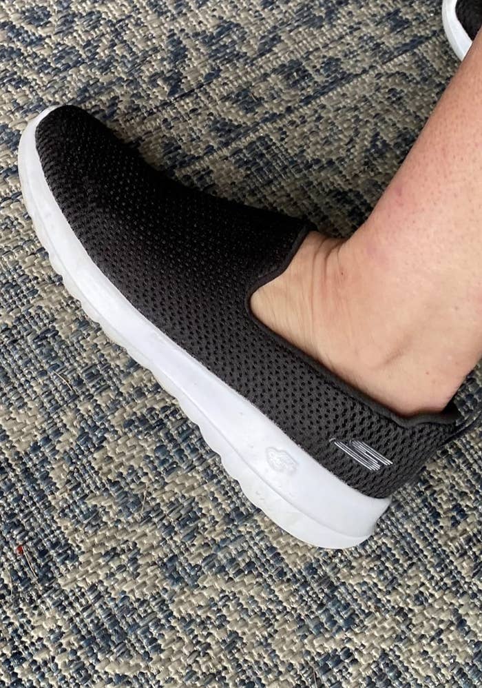 Close-up of a foot wearing a black mesh slip-on Skechers shoe with a white sole on a textured rug