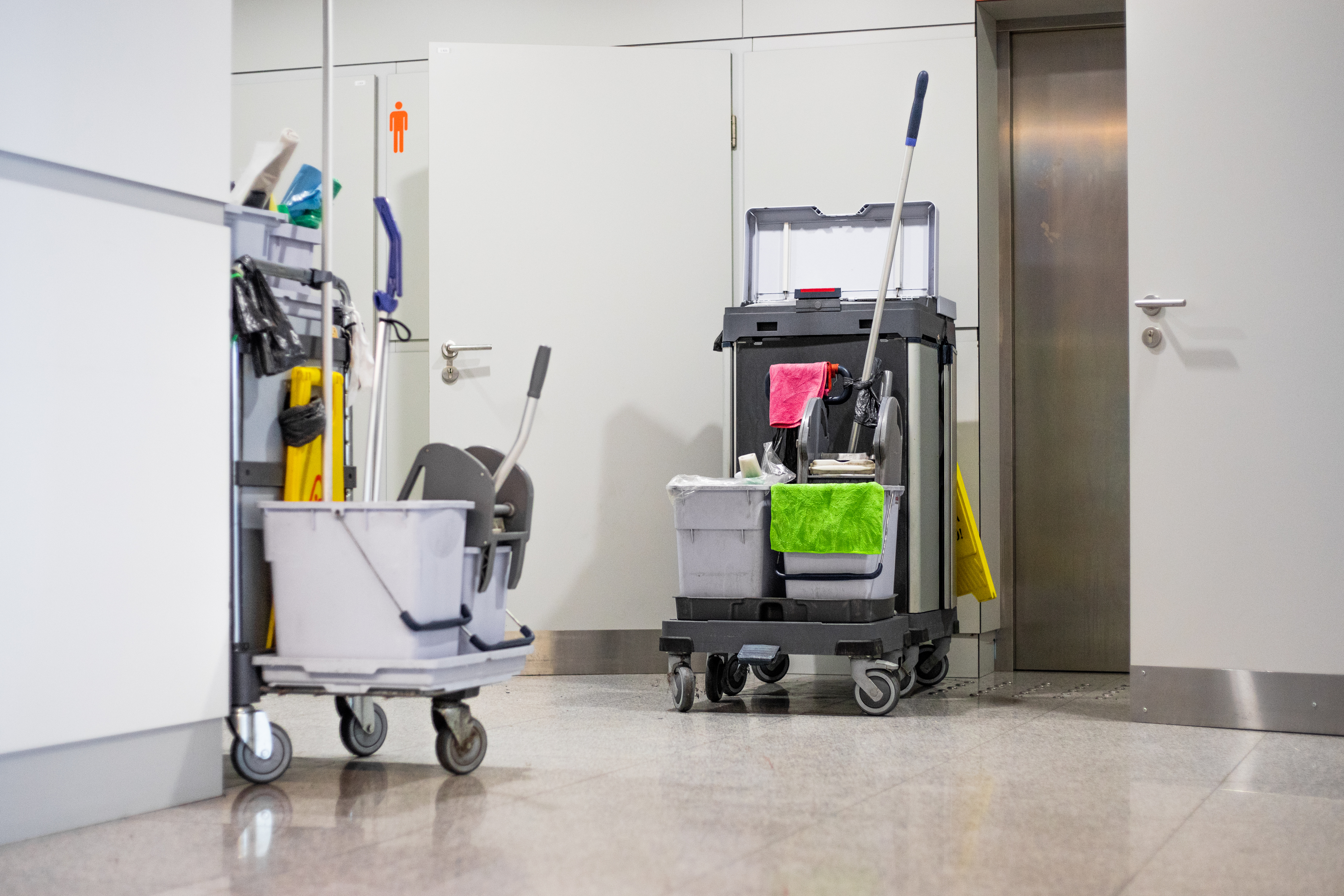 A janitorial station with two cleaning carts and various cleaning supplies near an open door and an elevator in a hallway