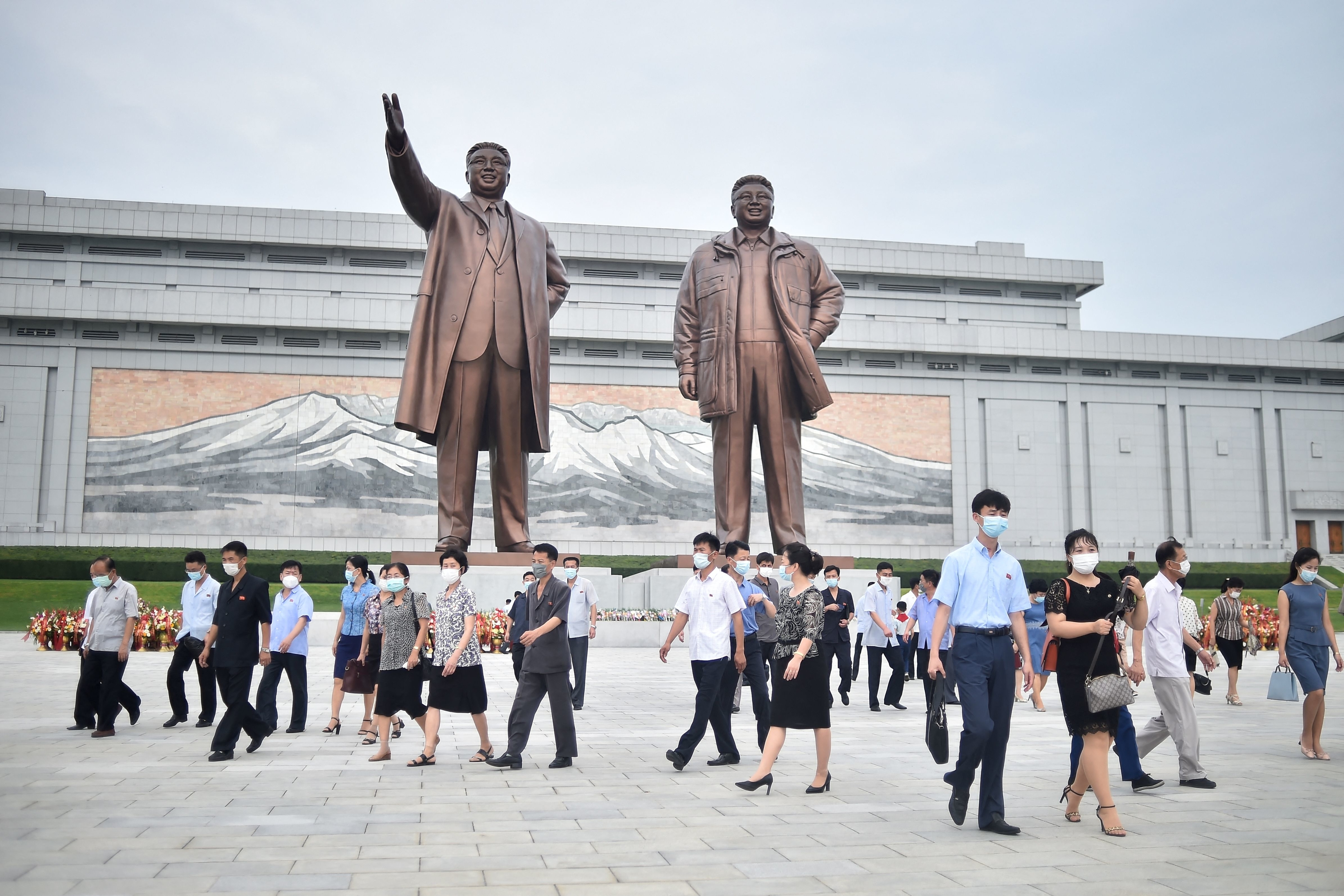 Statues of Kim Il-sung and Kim Jong-il stand tall while masked and socially-distanced people walk around in front of them at the Mansu Hill Grand Monument on July 27, 2022