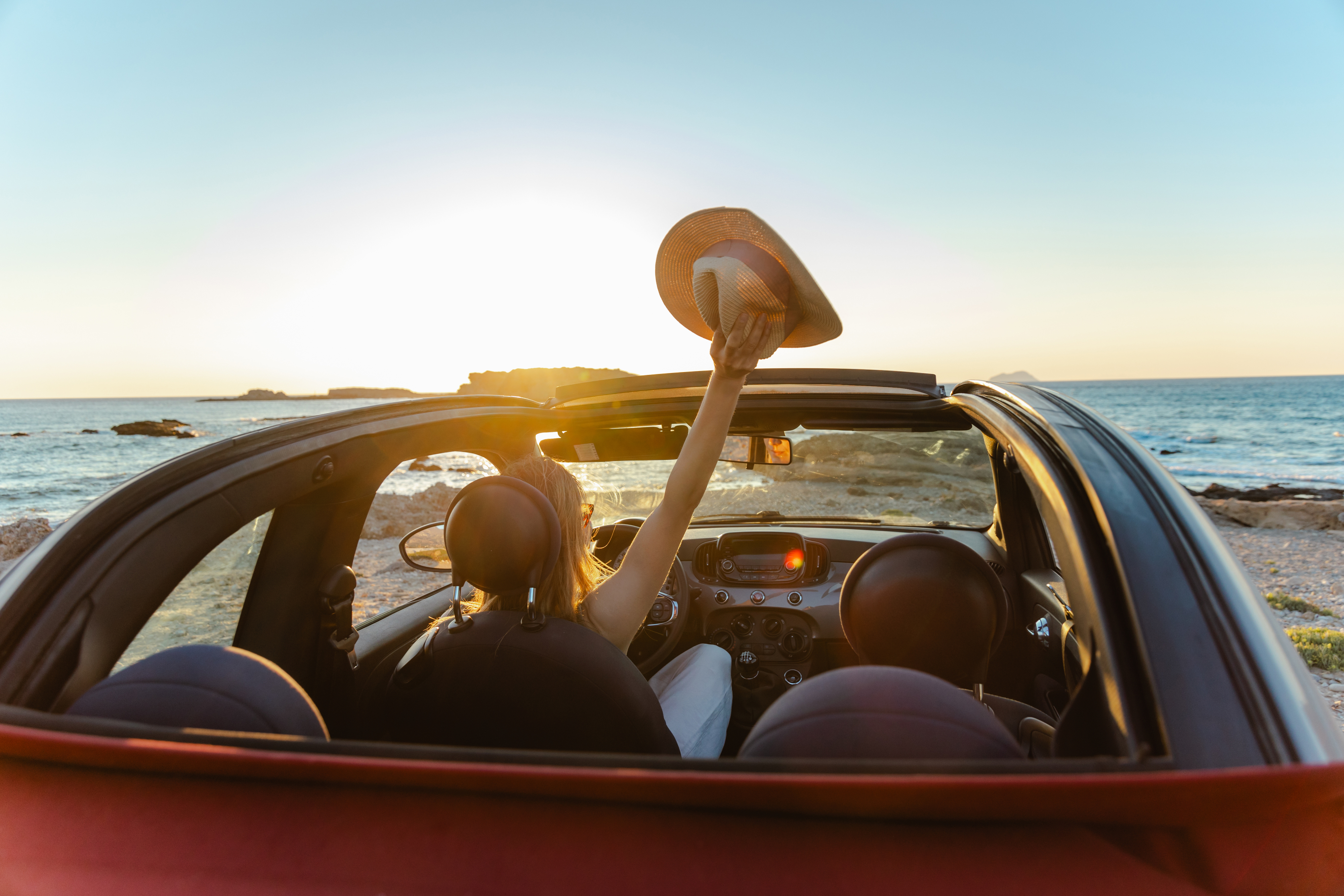 Rear view of a couple in a convertible by the ocean at sunset, one person holding a hat up