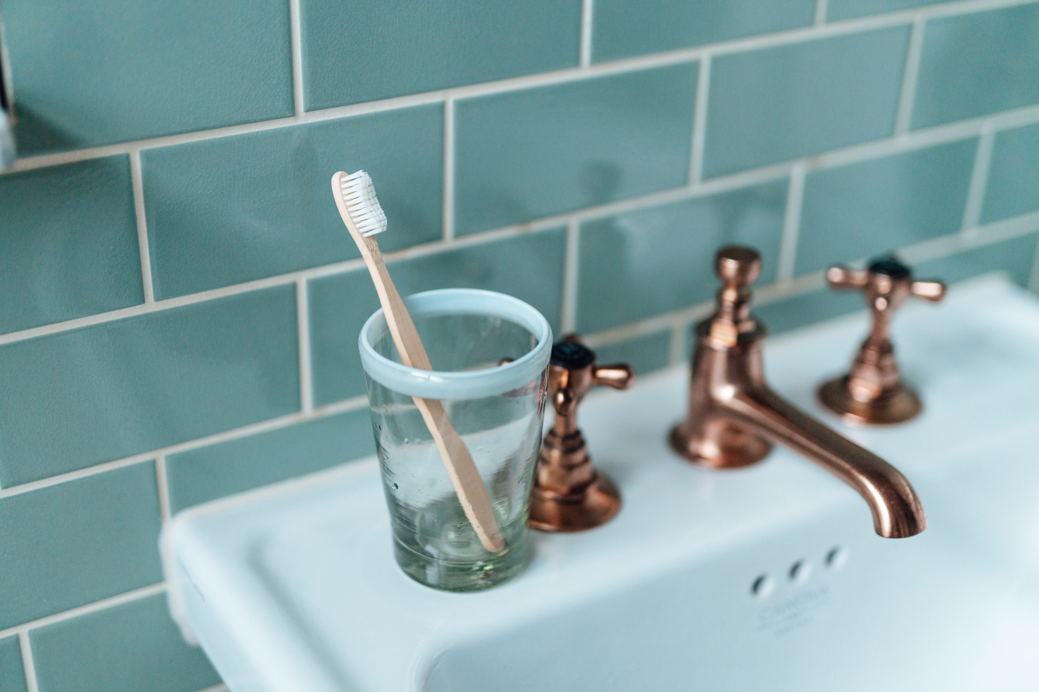A toothbrush is placed in a glass on a bathroom sink with brass faucets and a tiled backsplash