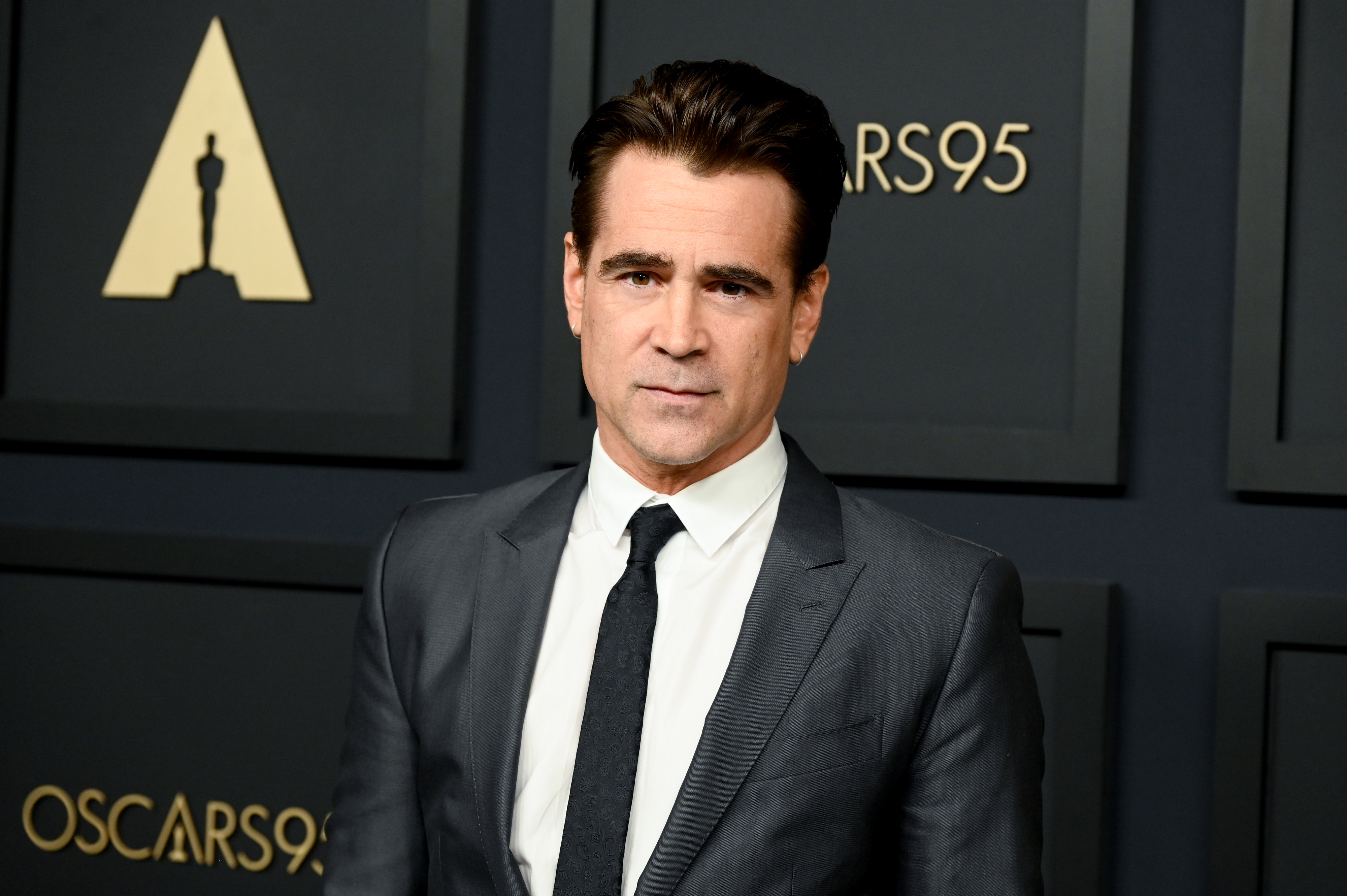 Colin Farrell in a formal suit and tie at the Oscars 95 event, standing in front of a black backdrop with Oscars logos