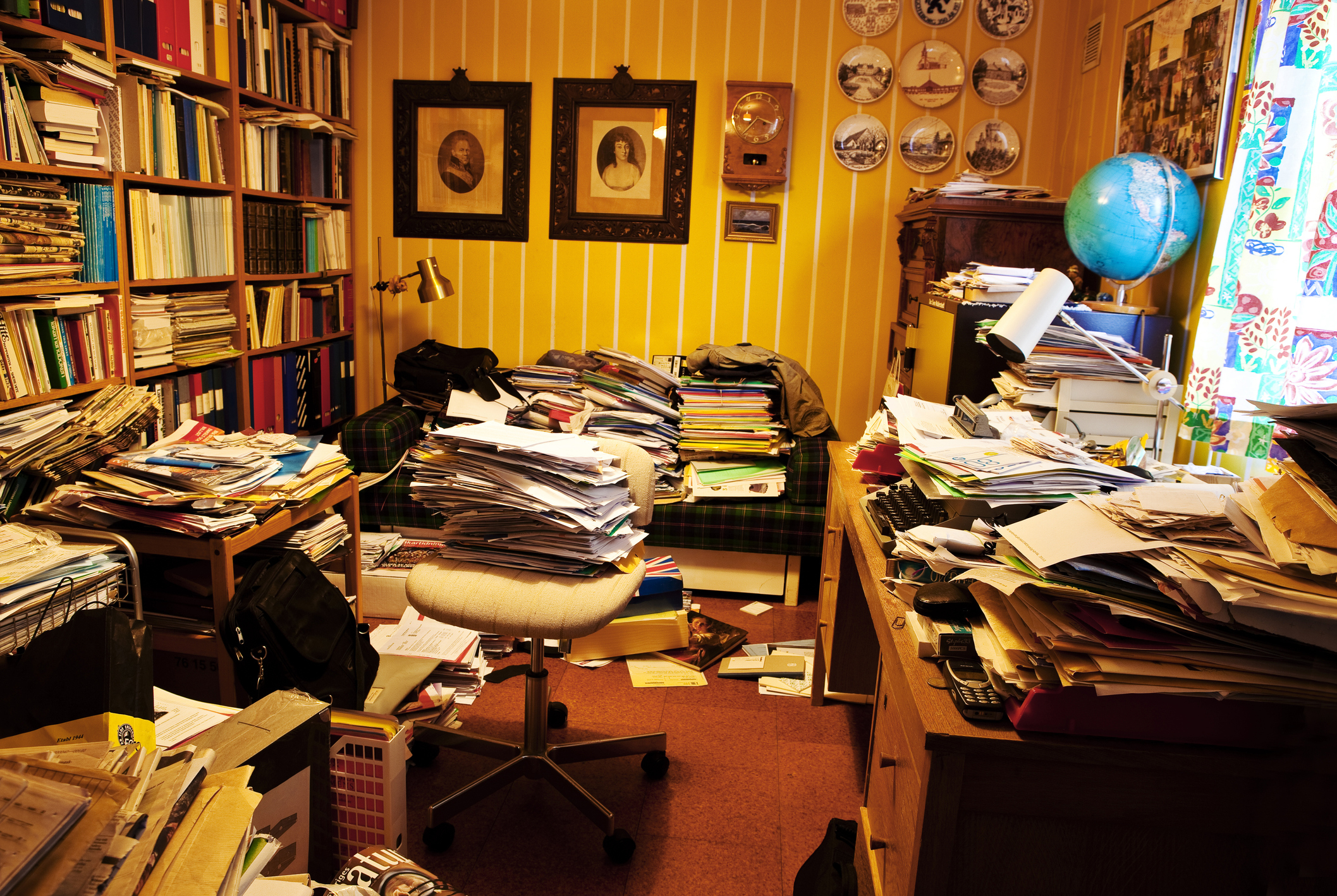 Crowded home office with stacks of papers, bookshelves, framed photos, and a globe on the desk. A chair is centered amid the cluttered workspace