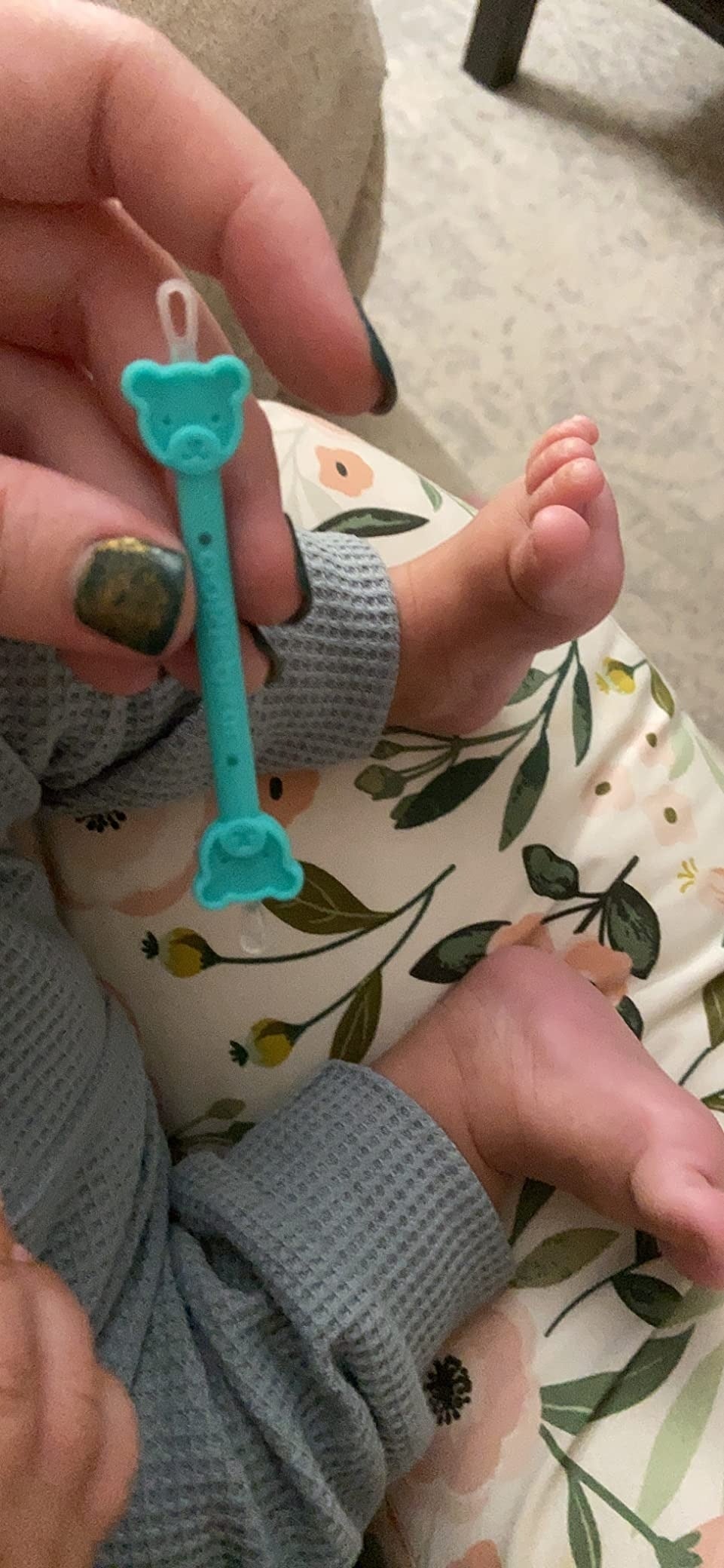 Hand holding a bluish  babe  teething artifact  shaped similar  a bear, adjacent   a baby's feet. The babe  is wearing a waffle-textured outfit