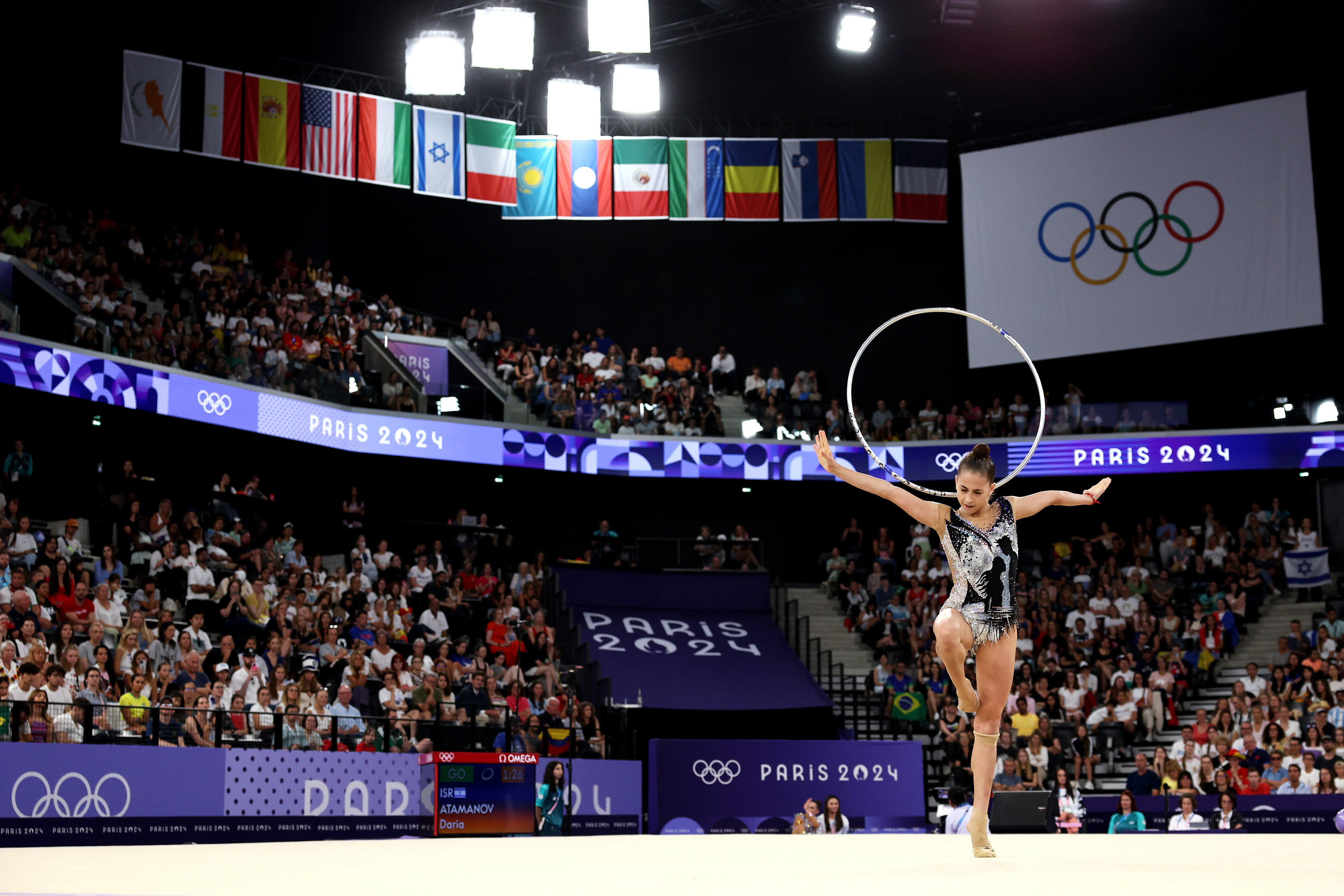 A gymnast performs with a hoop at the Paris 2024 Olympics. The arena is filled with spectators and country flags. The Olympic rings are displayed on a screen