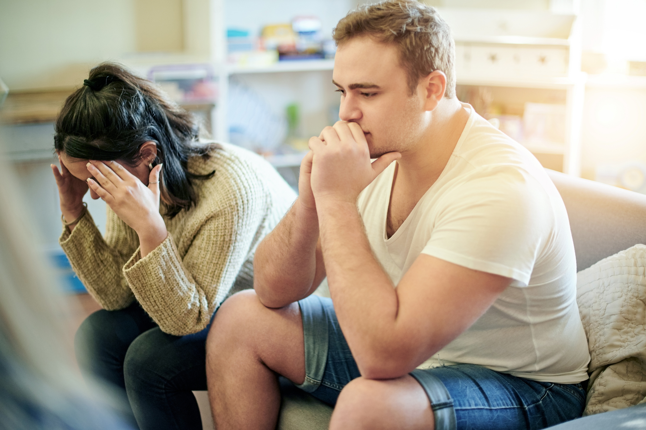 A man and a woman sitting next to each other on a couch, both look distressed; she has her head in her hands, and he is leaning forward with his hands near his mouth