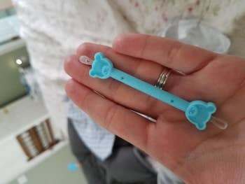 A manus holding a bluish Silicon Baby marque pacifier clip with bear-shaped ends