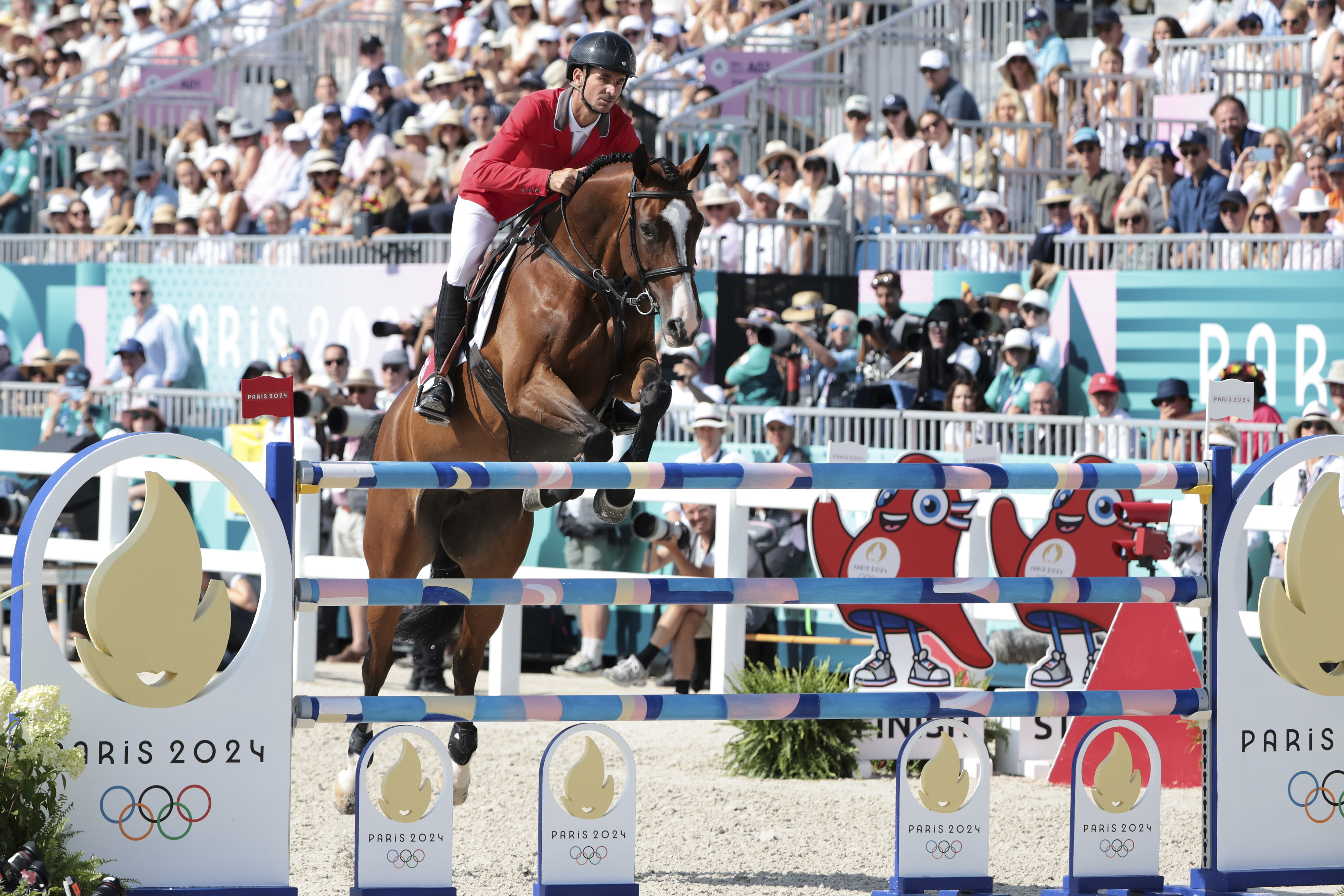 An equestrian rider, wearing a helmet and red jacket, jumps over an obstacle with a brown horse during the Paris 2024 Olympic event. Crowd watches behind