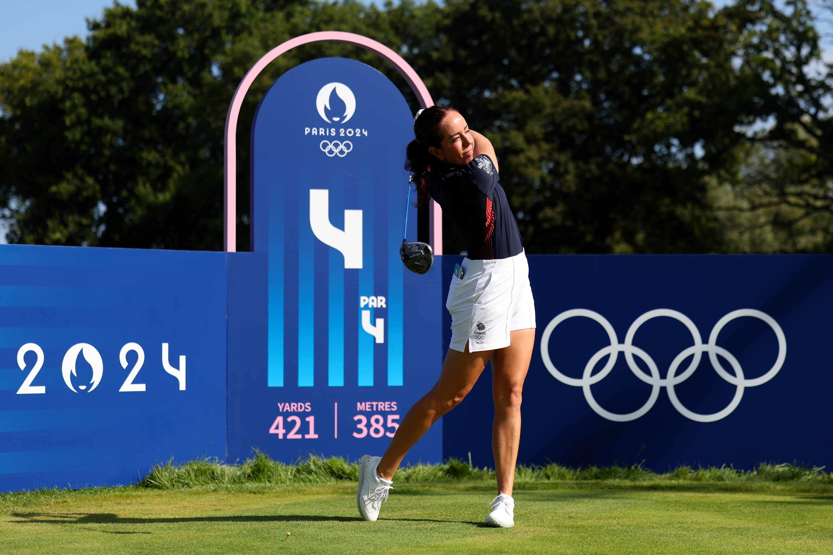 Golfer hitting a tee shot at the Paris 2024 Olympics golf course. The sign in the background reads "Par 4, 421 Yards, 385 Metres."