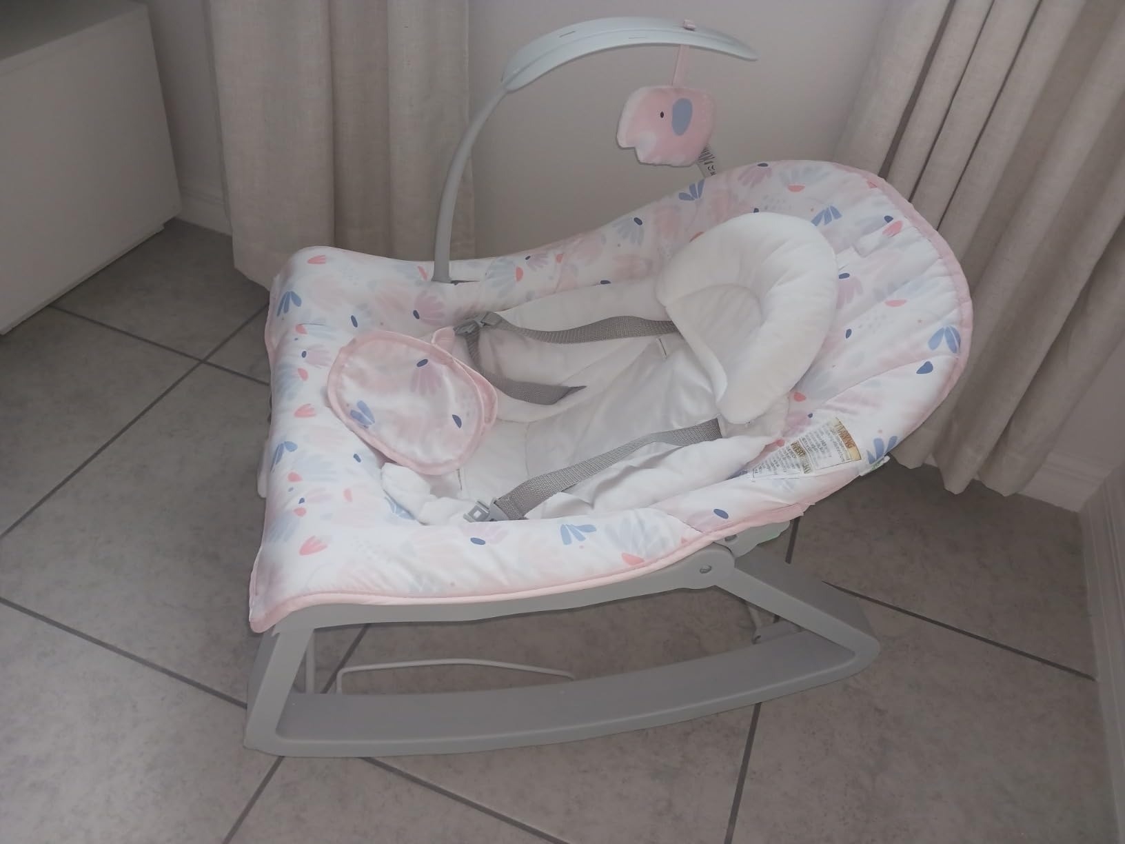 Baby bouncer seat with cushioned head support and safety straps. A mobile with two hanging soft elephant toys is suspended above